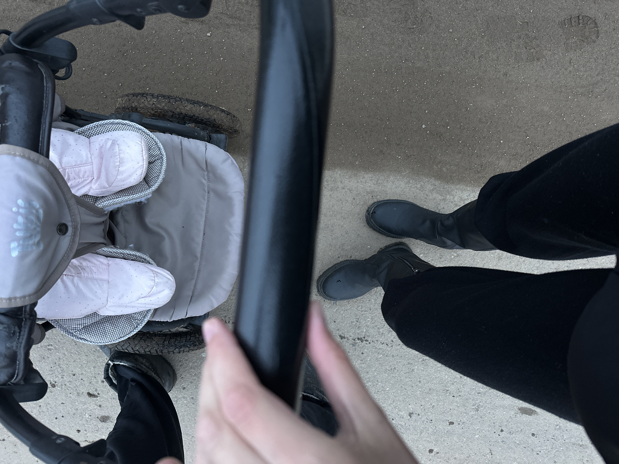 The image depicts a first-person perspective of someone pushing a stroller on a concrete surface. The stroller contains a baby wearing pink booties. The person pushing the stroller is wearing black pants and black boots. The scene appears to be outdoors. The location, Подшивалово, Russia, is not discernible from the visual information provided.