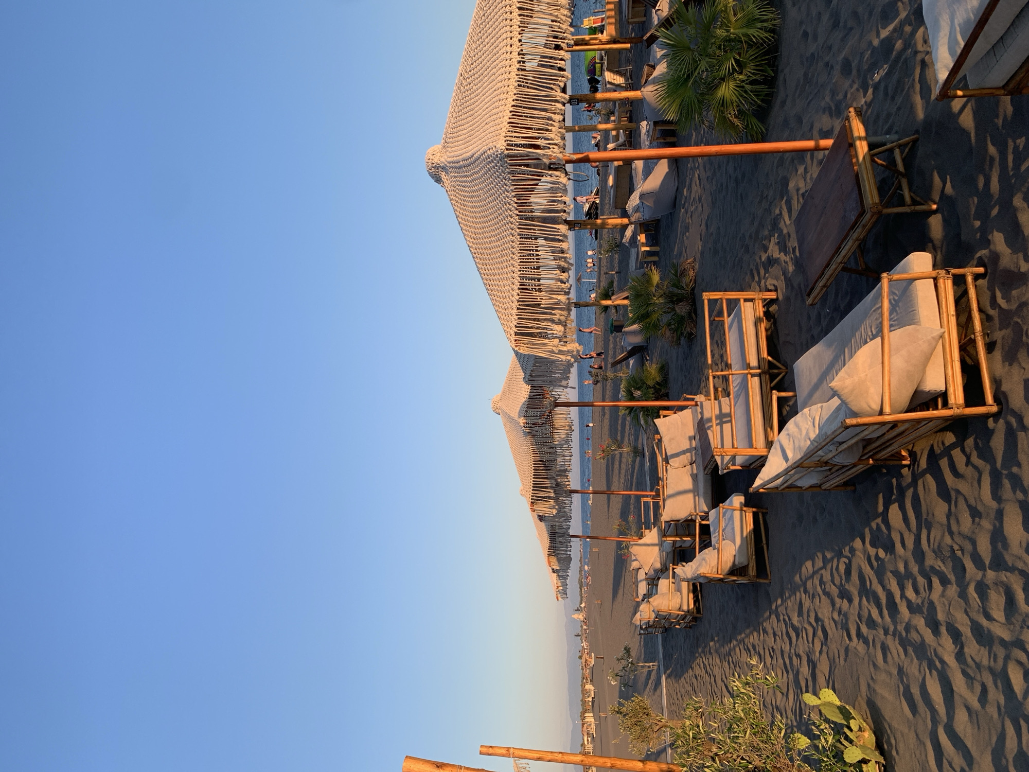 A beach scene at sunset in Donji Štoj - Shtoji i Poshtëm, Montenegro, features rows of bamboo-framed lounge furniture with light-colored cushions and woven umbrellas with fringe detailing. The dark sand is marked by shadows cast by the furniture and vegetation, including a cactus and palm plants. In the background, the sea is visible with several people walking along the shoreline and in the water. The sky is clear and blue, transitioning to a warmer hue near the horizon. Some wooden posts with string decorations are also present.