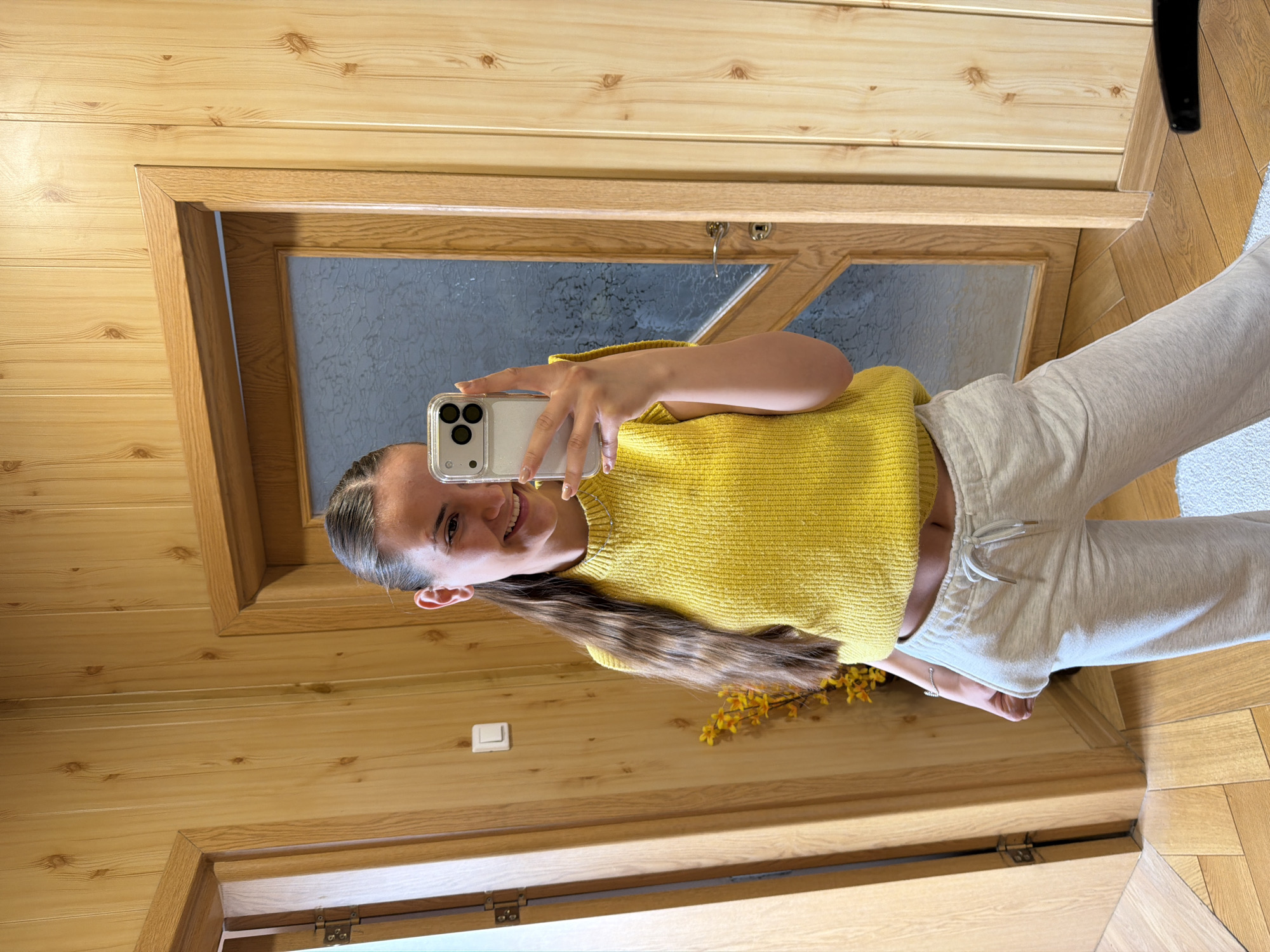 A young woman is taking a selfie in front of a wood-paneled wall and a wooden-framed window/door. She is wearing a yellow sweater and light-colored sweatpants. Her hair is pulled back, and she is smiling. Yellow flowers are near her waist. The location is possibly Buhovo, Bulgaria.