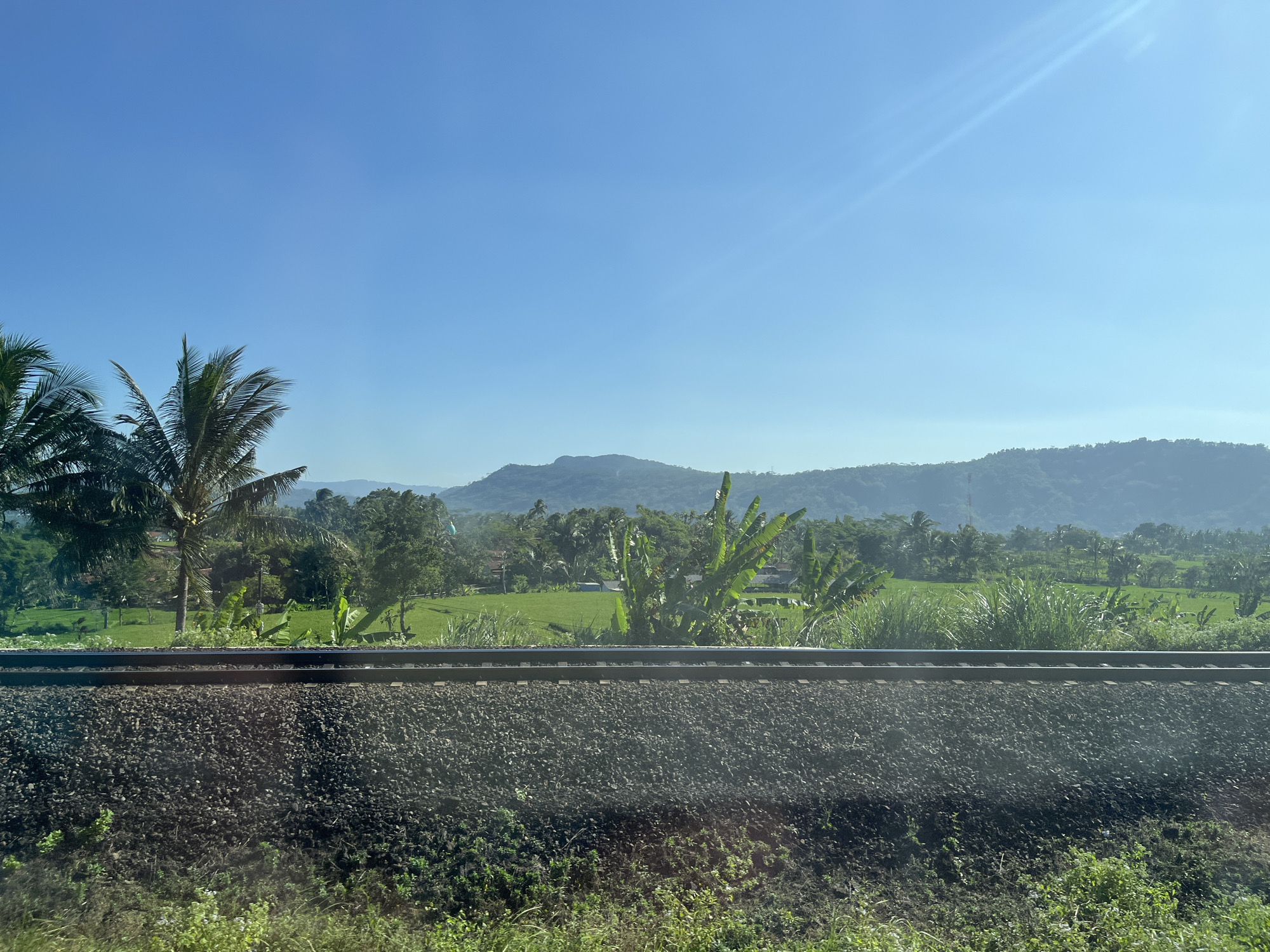 The image depicts a railway track in the foreground, with ballast and vegetation. Beyond the track, a lush green landscape unfolds, featuring rice paddies, palm trees, and other tropical vegetation. In the background, rolling hills are visible under a clear blue sky with a subtle sun flare. The scene is located in the city of Pasiraman Kidul, Indonesia. There are no people or animals visible in the image. The overall impression is one of a serene rural landscape.
