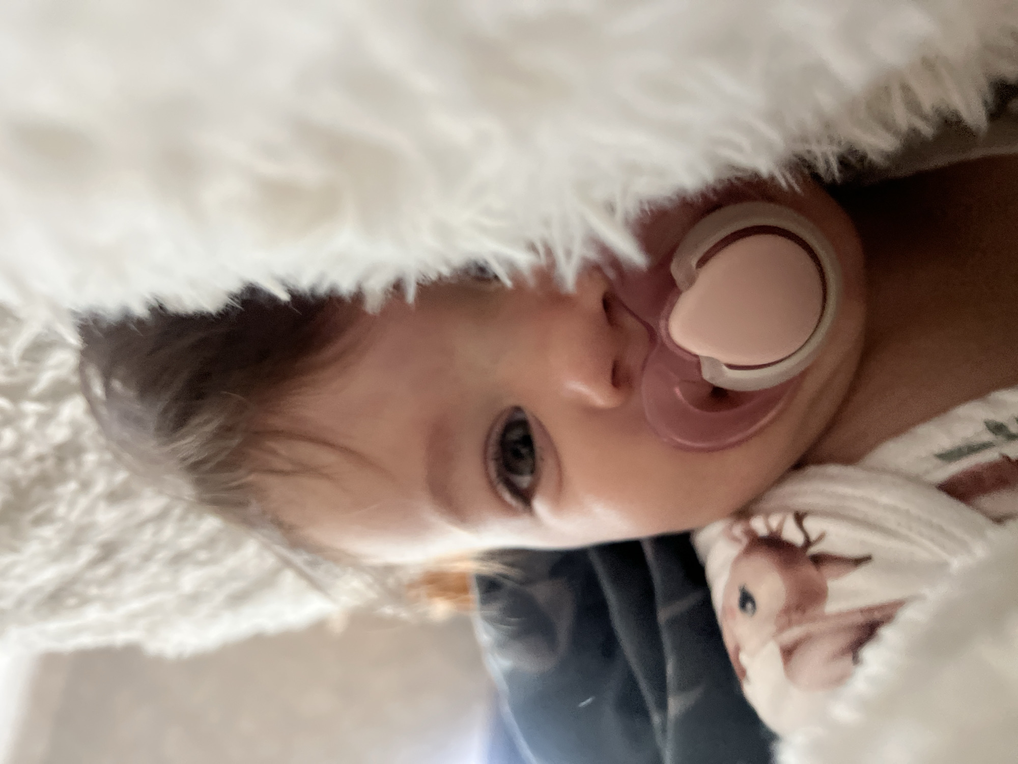 A young child with brown hair is lying down, possibly in a bed, with a pacifier in their mouth. The child is partially covered by a white, fluffy blanket. The child is wearing clothing with a deer pattern on it. The background is indistinct. The location is not discernible from the image.