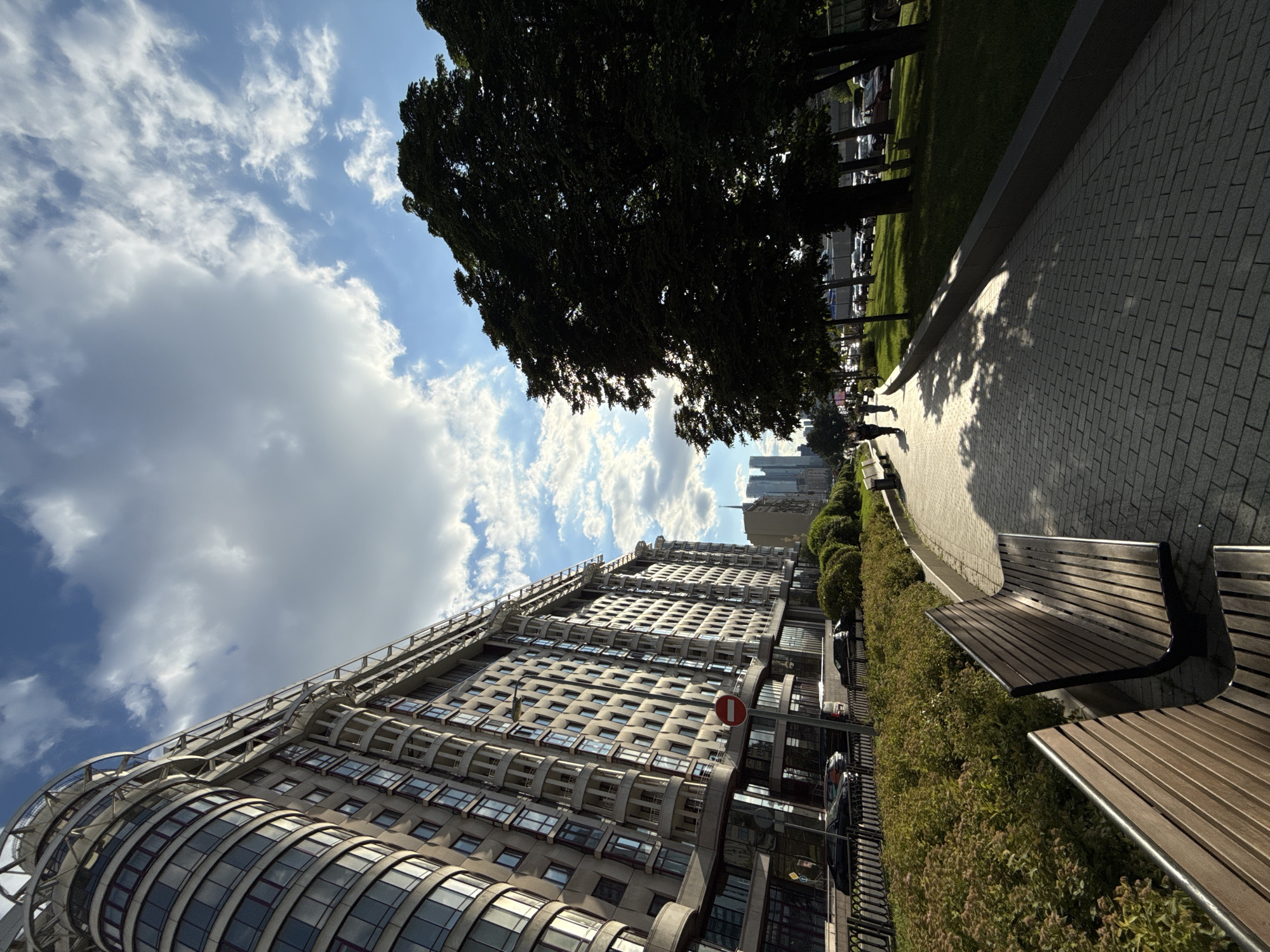 The image depicts a sunlit urban scene with a prominent architectural element and natural surroundings. A tall, modern building with a curved facade, featuring numerous windows and balconies, dominates the left side of the frame. Its reflective surfaces mirror the bright blue sky filled with scattered white clouds, suggesting fair weather.
To the right of the building, a paved walkway, likely made of stone tiles, stretches into the distance, casting dappled shadows from overhead trees. A park-like area with green grass and mature trees borders the walkway, and a row of wooden benches is positioned along the edge, indicating a public space designed for relaxation. In the background, other buildings, including skyscrapers, are visible, hinting at a city environment.
A few people are visible walking on the pathway, appearing as small figures against the vastness of the architecture and landscape. The time of day is likely midday or early afternoon, given the strong sunlight and pronounced shadows. The overall mood is serene and vibrant, highlighting the interplay of modern construction and green spaces. No legible text is clearly visible within the image.