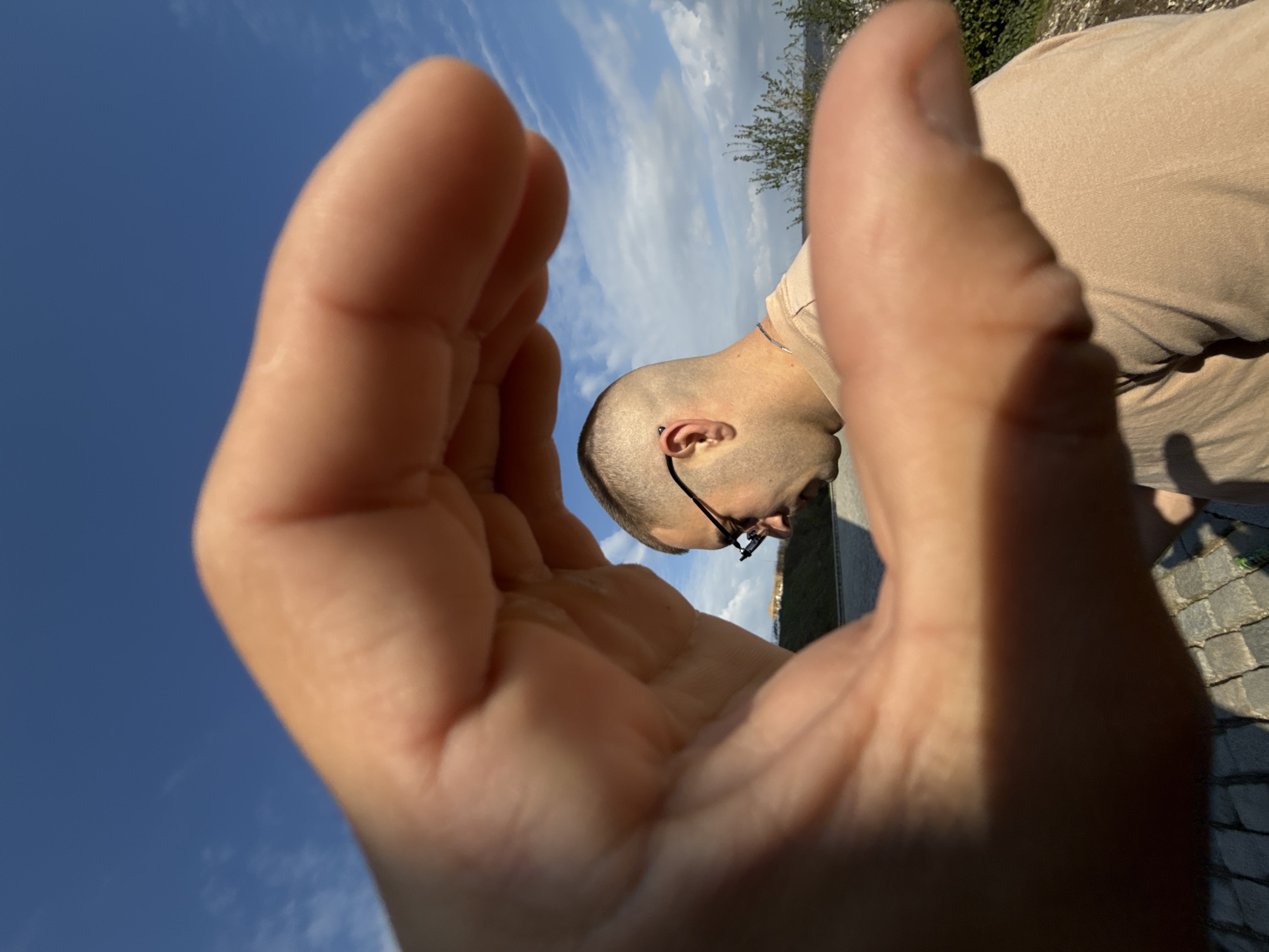 A man stands outside in daylight, close to a paved surface. The man's head and upper torso are visible. He has a shaved head, and wears glasses. The man is wearing a light beige shirt. He appears to be looking downwards. The camera angle is unusual, featuring a hand in the foreground, partially obscuring the man and the background. The sky is blue with some white clouds. In the distance are some green trees and a body of water. The location is plausibly Shumen, Bulgaria.