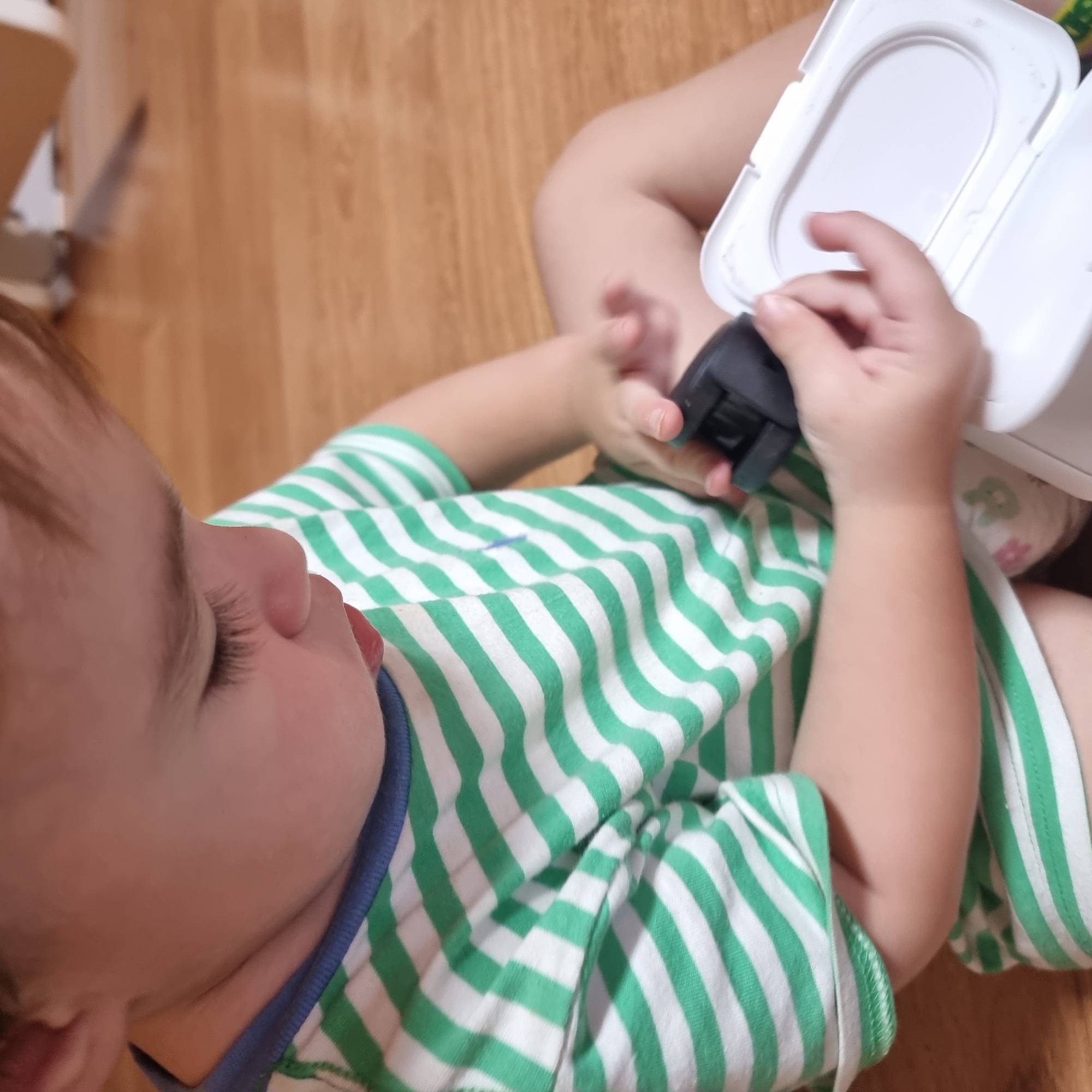 A toddler is sitting on a wooden floor and playing with a wet wipe container. The toddler is wearing a green and white striped t-shirt and a diaper with a bunny print. The toddler's hands are in focus as they interact with the white container and a black object. The background is blurred and shows more of the wooden floor and a hint of furniture. The image is taken from a close-up perspective, focusing on the toddler's engagement with the objects.