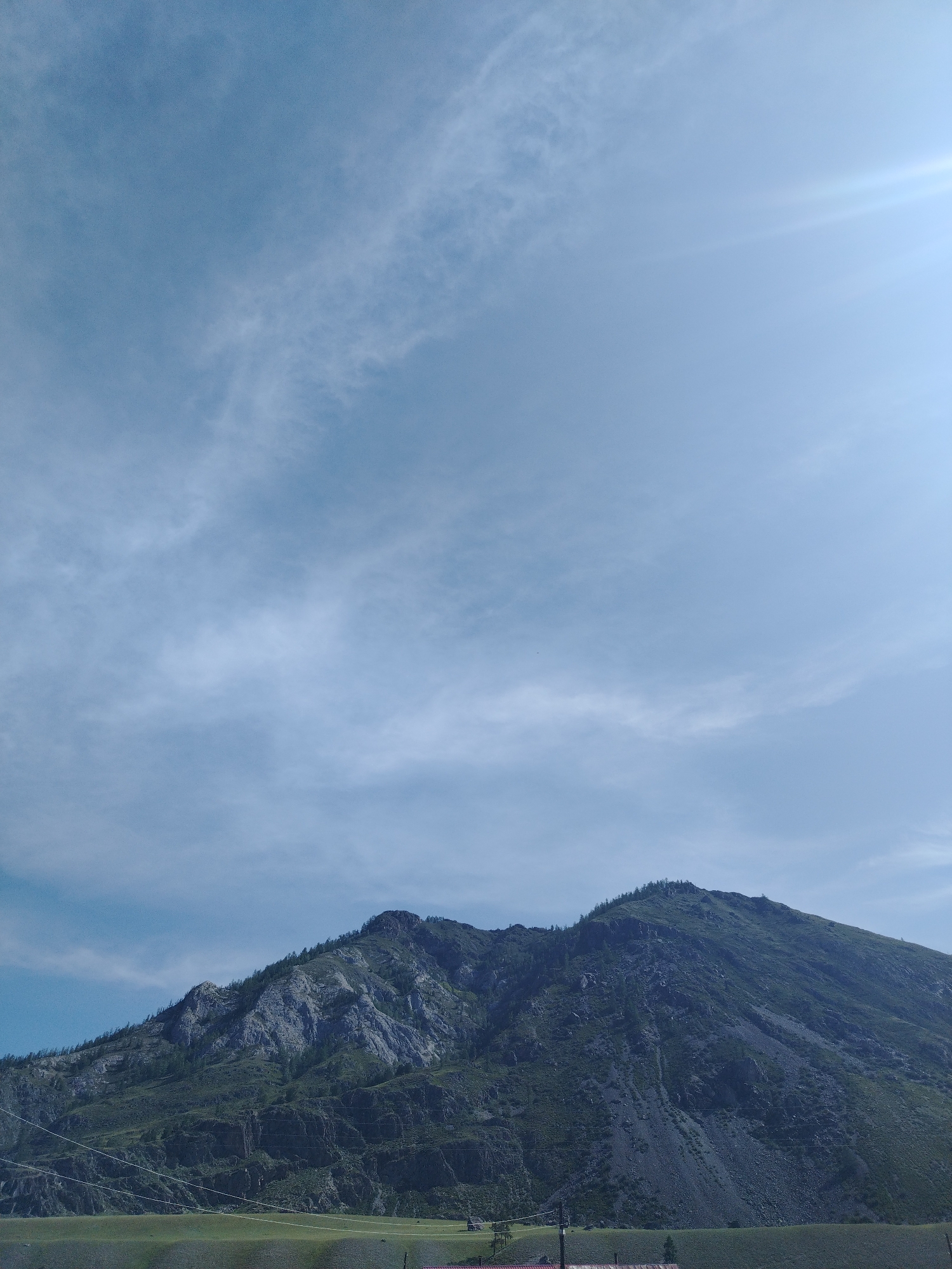 The image shows a mountainous landscape under a partly cloudy sky. The foreground features a grassy field with some distant structures. A prominent, steep mountain with rocky outcrops and scattered trees dominates the middle ground and background. Power lines are visible in the lower part of the mountain. The sky is a pale blue with wispy clouds and a bright sunbeam in the upper right corner. The scene appears to be a natural, outdoor environment, possibly in a rural or mountainous region of Russia, such as Iodro, known for its natural beauty.