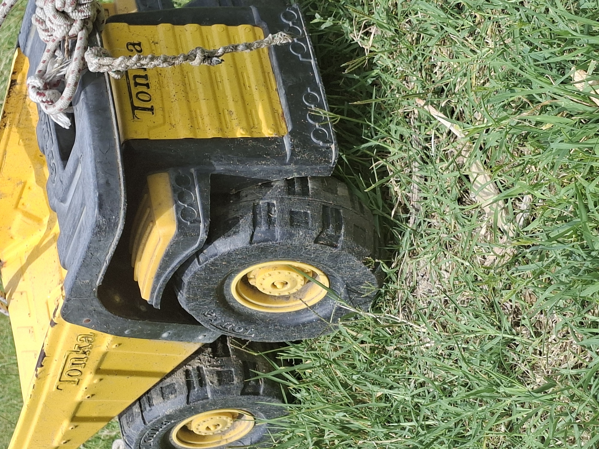 A close-up, eye-level shot shows a bright yellow toy dump truck, a Tonka truck, resting on green grass. The truck is partially submerged in the grass, with its large black tires and yellow rims visible. The truck has the "Tonka" logo embossed on its side. A thick, white and red rope is loosely draped over the truck's black plastic hood. The scene is outdoors and appears to be a sunny day. The image does not provide any information that would allow for its location to be identified within the city of Tbilisi, Georgia, or any other specific location.