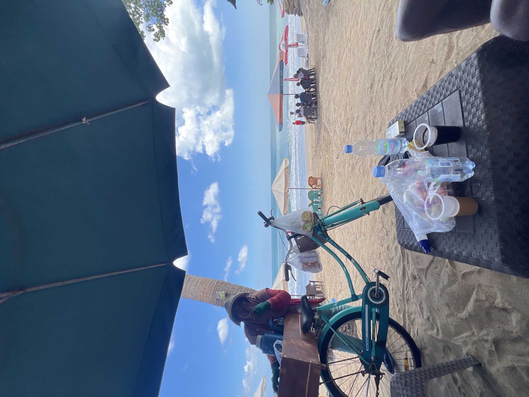 A beach vendor with a turquoise bicycle is seen on the sand in Kuta, Indonesia. The vendor wears a hat, gloves, and a red jacket over a pink shirt, and is positioned behind the bicycle, which has a wooden cart attached. Two plastic bags filled with items are hanging from the bicycle handlebars. In the foreground, a dark plastic table holds two bottles of water, a pack of cigarettes, and two cups, one with a dark liquid and the other with a lighter brown liquid. The beach is lined with umbrellas and lounge chairs, and several people are seated under a large umbrella in the background. The ocean and a cloudy blue sky are visible.