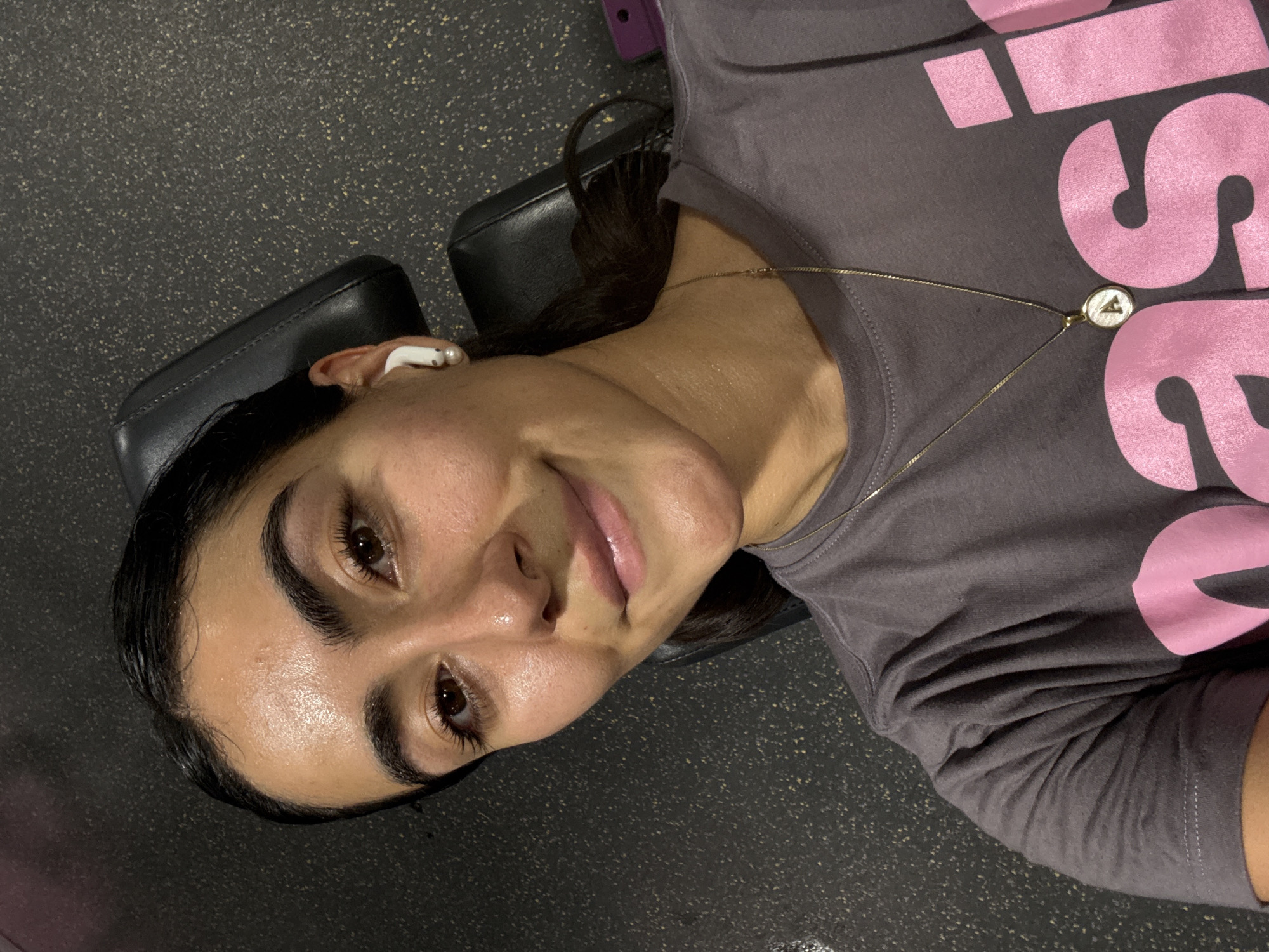 A woman with dark hair, wearing a gray t-shirt with pink lettering and a gold necklace with a circular pendant bearing the letter "A", lies on a padded black bench with an in-ear headphone in her left ear. The background consists of a speckled gray floor. The location is a gym. The specific location in Puebla, Mexico is indeterminable without further information.