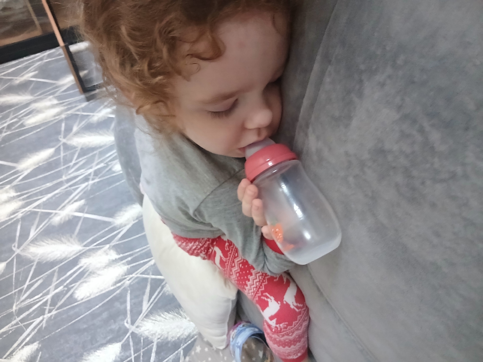 A young child, likely a toddler, with fair skin and curly reddish-brown hair, is captured in a close-up, top-down shot. The child's eyes are closed, indicating they are either asleep or deeply drowsy, leaning against a large, soft, gray upholstered surface, possibly a sofa or cushion. They are holding a clear plastic baby bottle with a pink cap and nipple, which appears to contain a small amount of liquid.
The child is wearing a light gray long-sleeved top, light-colored pants, and distinctive red leggings or socks patterned with white reindeer and snowflake motifs, suggesting a festive or winter theme. To the left of the child, the floor is covered with a gray rug or carpet featuring a white, abstract linear and feather-like pattern. A dark wooden or metal frame is partially visible in the upper left corner of the background, hinting at nearby furniture or room decor. The overall scene conveys a sense of peaceful comfort and rest. No visible text is present in the image.