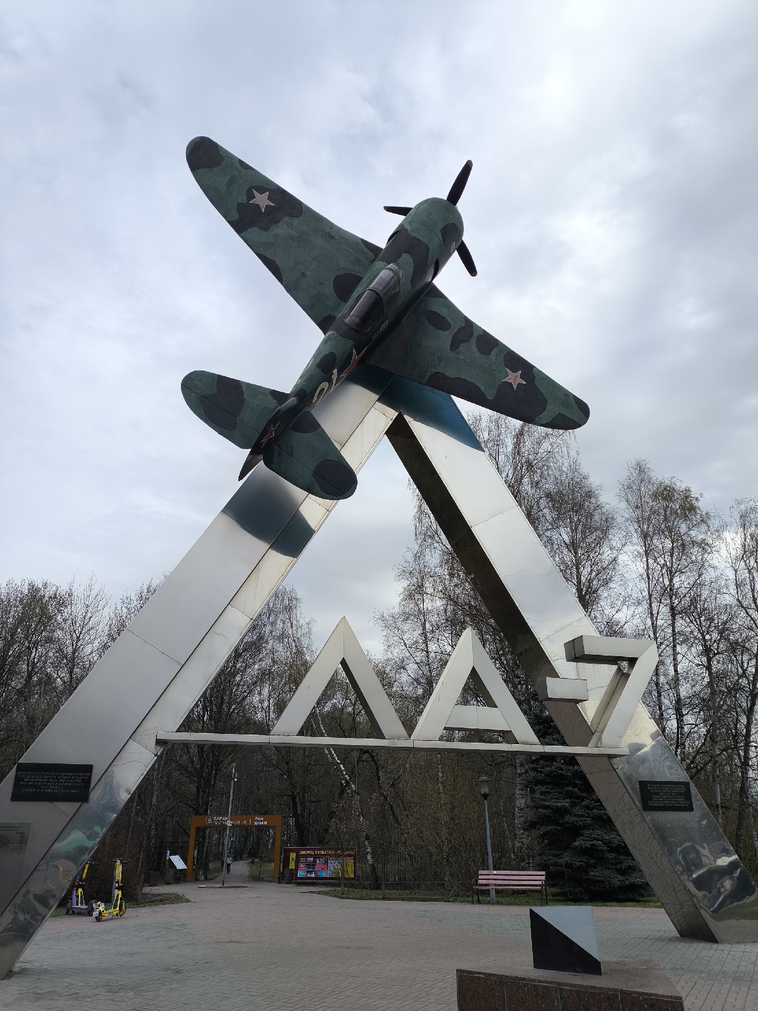 A large monument featuring a Soviet-era fighter plane, likely a Lavochkin La-7, stands prominently under an overcast sky in Khimki, Russia. The aircraft, painted in a green and dark camouflage pattern with red stars on its wings and fuselage, is mounted on a striking metallic archway. The plane is angled upwards, as if taking off, with its propeller facing towards the upper left.
The monument's archway is composed of two large, gleaming metallic beams that converge at the top and form the letters "ЛА-7" (LA-7), the designation for the aircraft model. On the left support of the monument, a plaque with Cyrillic text is visible. It states that the monument was erected by the administration of the Khimki city district in memory of the first batch of La-7 fighters produced at Khimki Plant No. 301, with construction completed in 2007. A similar plaque appears on the right support, likely providing further details about the aircraft or its historical significance.
The monument is set in a paved area, suggesting a public park or square. In the background, numerous deciduous trees with bare branches and some evergreen trees are visible. Through the archway, a brown wooden archway marking the entrance to a park can be seen, with Cyrillic text that reads "ПАРК КУЛЬТУРЫ И ОТДЫХА ДУБКИ" (Park of Culture and Recreation Dubki). To the right of this entrance, another sign indicates "ДВОРЕЦ КУЛЬТУРЫ" (Palace of Culture).
In the foreground, near the left support, two yellow and black electric scooters are parked. A red wooden bench is positioned near the right support, and a dark, geometric ground sculpture sits in front of it. The overall impression is that of a historical memorial in a community park, presented during daytime under a cloudy sky.
