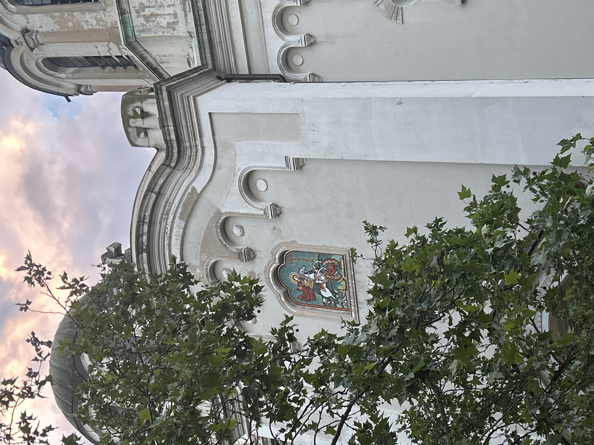 A low-angle, vertically oriented photograph captures a segment of a historic building, likely a church in Sofia, Bulgaria, partially obscured by the vibrant green foliage of a large tree. The architecture features ornate details, including curved cornices, decorative pilasters, and rounded arch motifs on a light-colored, possibly stucco or stone, facade that exhibits signs of weathering and age. A weathered dome or tower, clad in what appears to be metal, is visible on the far left.
The focal point is a striking mosaic embedded in the wall, set within an ornate, multi-lobed niche. The mosaic depicts a dynamic scene of a haloed saint on horseback, valiantly spearing a dark, monstrous dragon. The saint is dressed in a red tunic and blue cloak. Above the saint's halo, the Cyrillic text "СВ.ГЕОРГI" (ST. GEORGI) is clearly visible, identifying the figure as Saint George. The mosaic is rendered in vibrant colors using small tesserae, showcasing intricate detail.
The sky in the upper background reveals soft hues of pink and purple within the clouds, indicative of either sunrise or sunset. The diffused light suggests a cloudy yet calm atmosphere. The image captures a blend of architectural history, religious art, and natural elements, conveying a sense of enduring cultural heritage. No specific human activity is depicted in the frame.