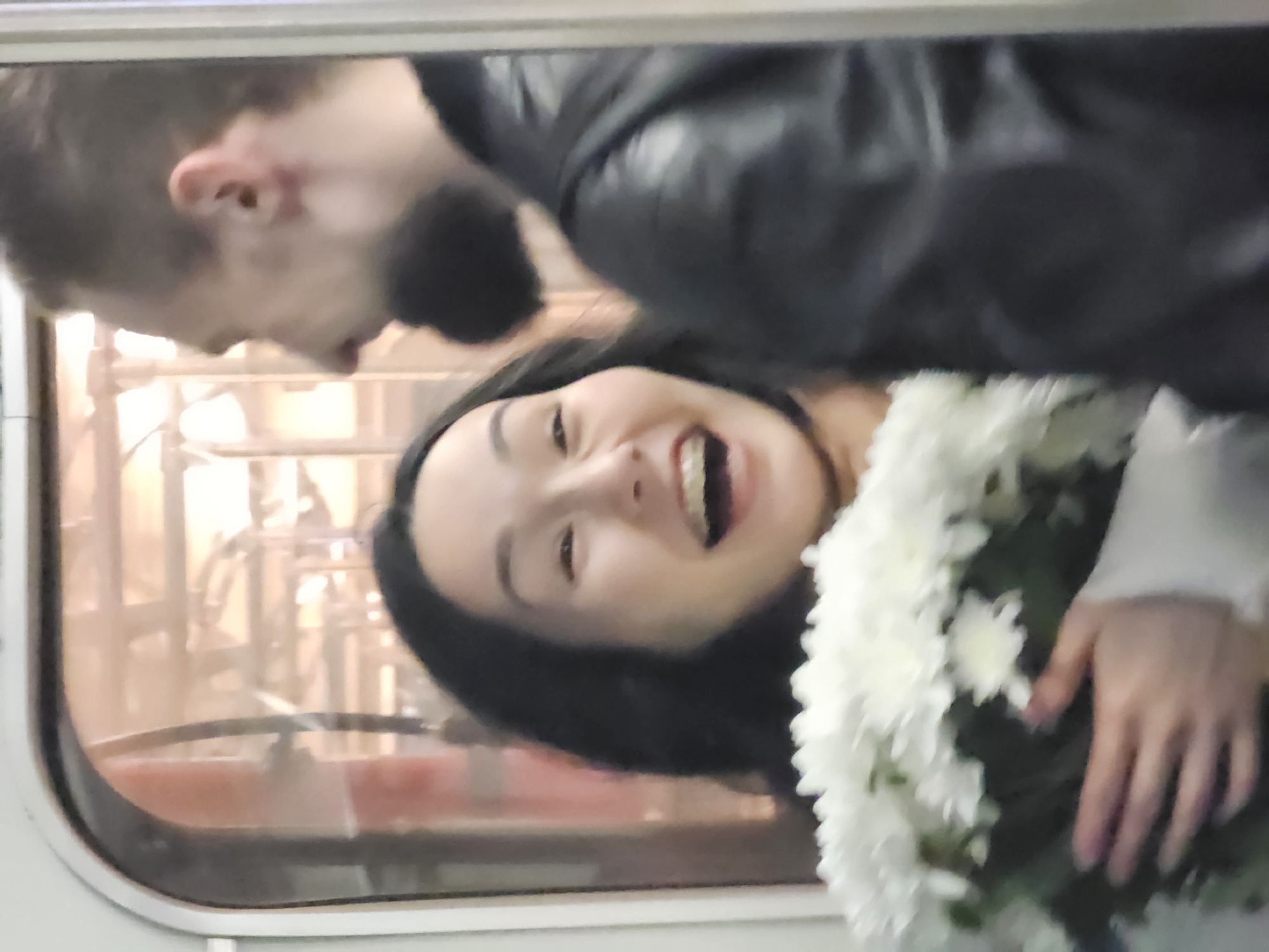 A woman with black hair is laughing while holding a bouquet of white flowers. A man with a beard is standing beside her, wearing a black leather jacket. They appear to be inside a train car, as indicated by the window and interior view. The location is Unknown, Unknown.