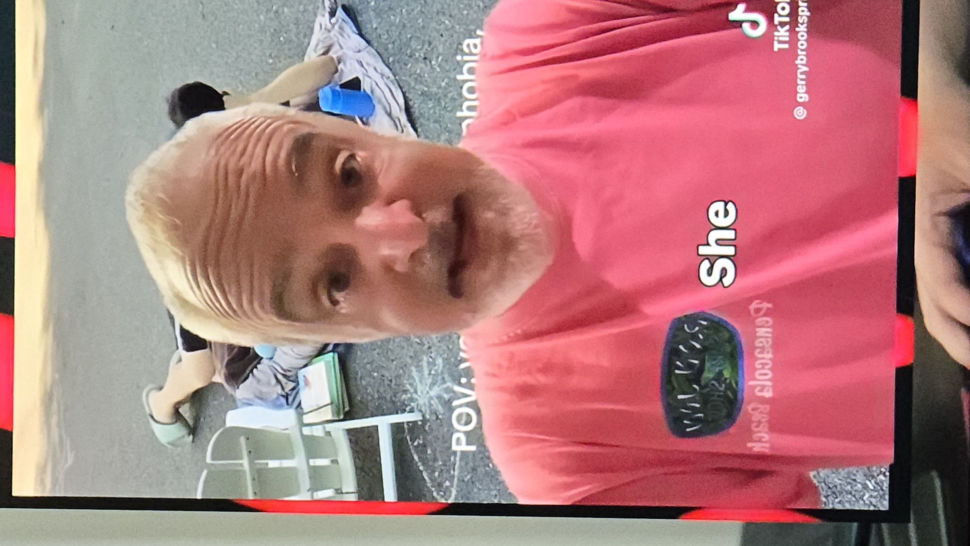 A close-up, first-person perspective shot shows a man with blonde hair and a pink t-shirt. He is in the foreground, with a blurred background. Behind him, a person lies on the ground, their legs bent and one foot in a light blue sandal. A white plastic chair is visible to the left of the person on the ground. Text overlays the image, with "POV:" and "phobia," visible in white lettering on the ground. A circular logo with green and black colors is on the man's t-shirt, and the word "She" appears in white text to the right of the logo. A TikTok logo and username "@gerrybrookspri" are in the bottom right corner. The overall environment appears to be outdoors, possibly a street or paved area.