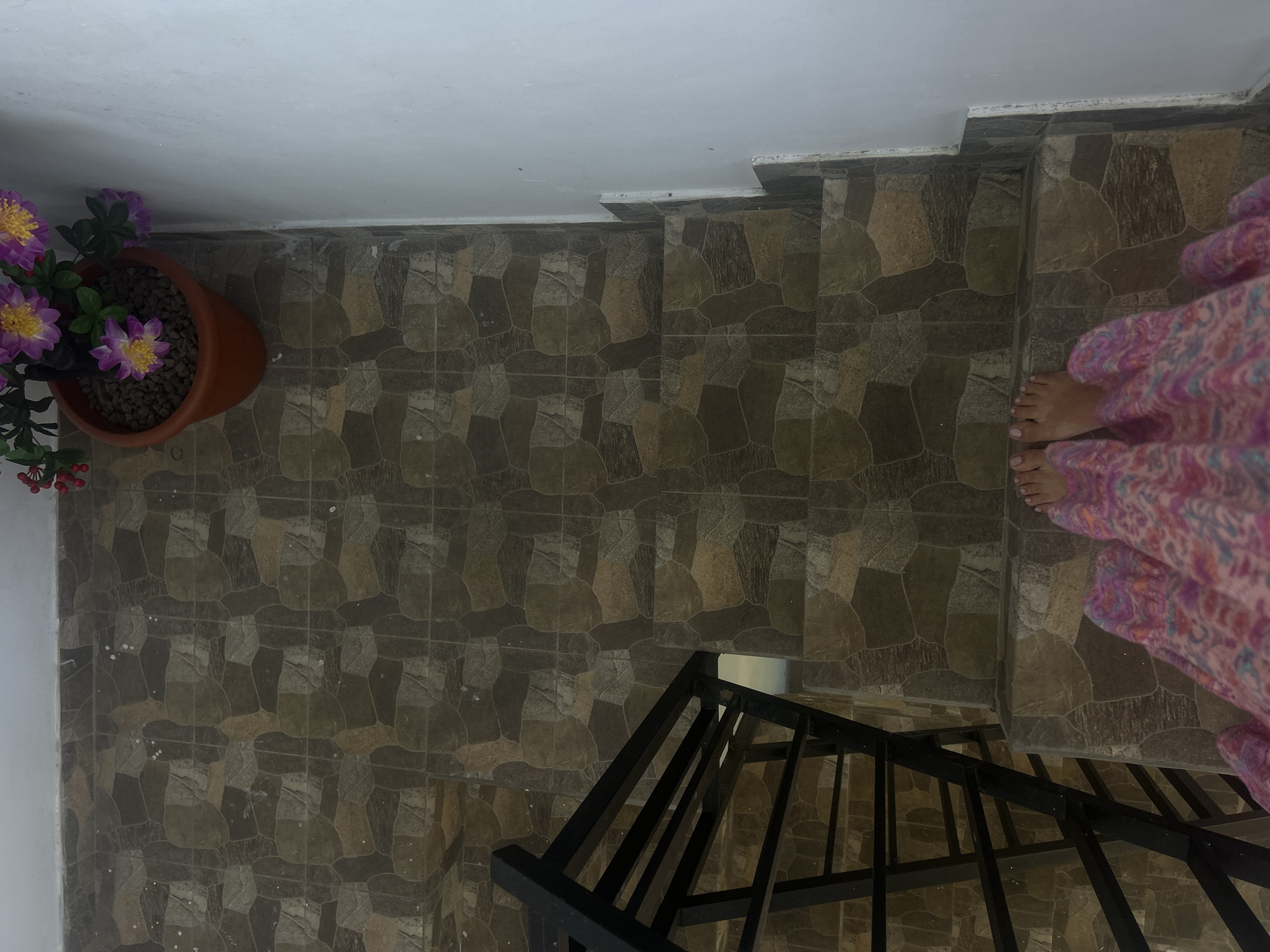 An overhead shot captures a person's feet and the lower part of their legs, adorned in a flowing pink and purple patterned garment, as they stand on a tiled staircase in North Kuta, Indonesia. The staircase features large, irregularly shaped tiles in shades of brown and beige, creating a mosaic-like pattern. A black metal railing is visible on the left side of the stairs. In the upper right corner, a terra cotta pot holds a faux purple and yellow flower arrangement, its small red berries adding a touch of color. The wall to the right of the staircase is painted white.