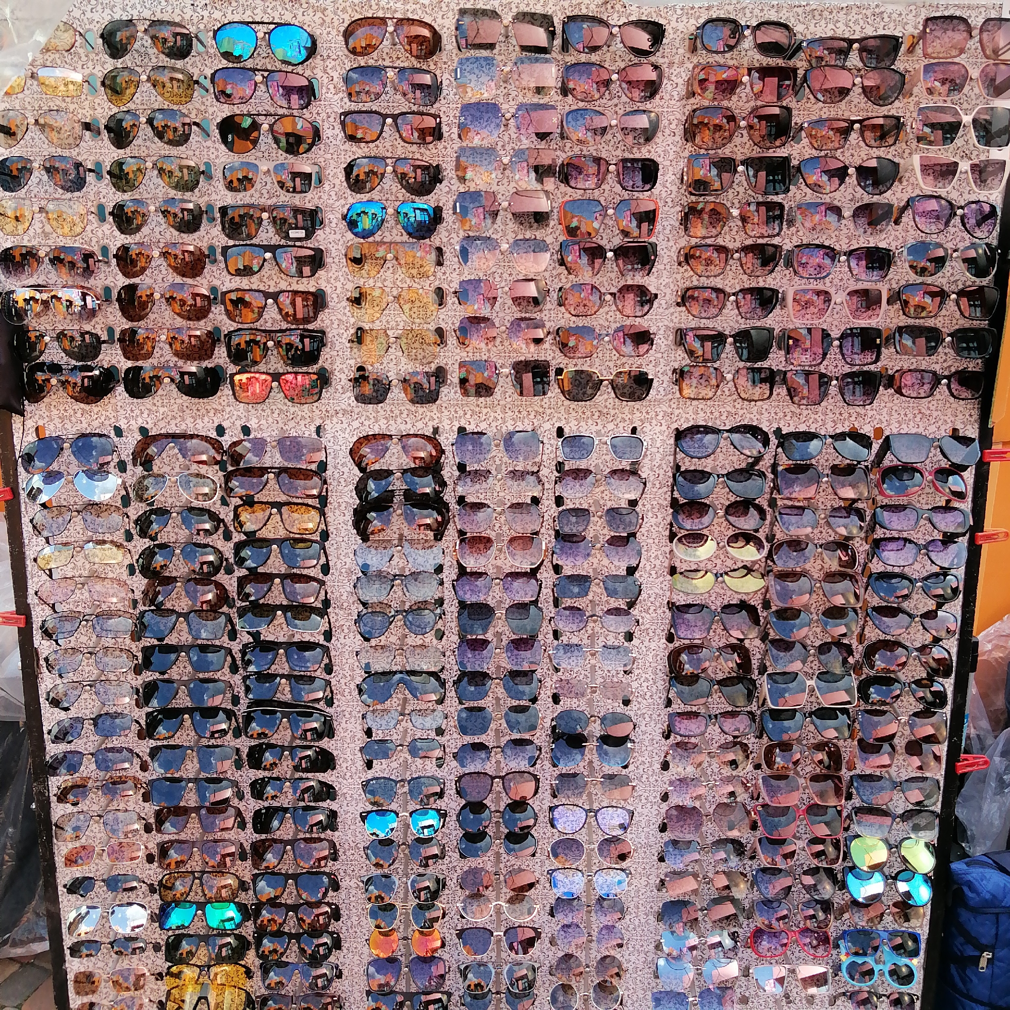 The image shows a large display of sunglasses arranged in rows and columns. The sunglasses have a variety of frame shapes and lens colors, including black, brown, blue, pink, and reflective lenses. The display is set up outdoors, likely at a market or street stall, as suggested by the presence of a blue bag and red clips visible on the right side of the image. The background is a patterned fabric, possibly a tablecloth or display cloth. The lighting suggests it is daytime, and the overall scene appears to be a retail environment.