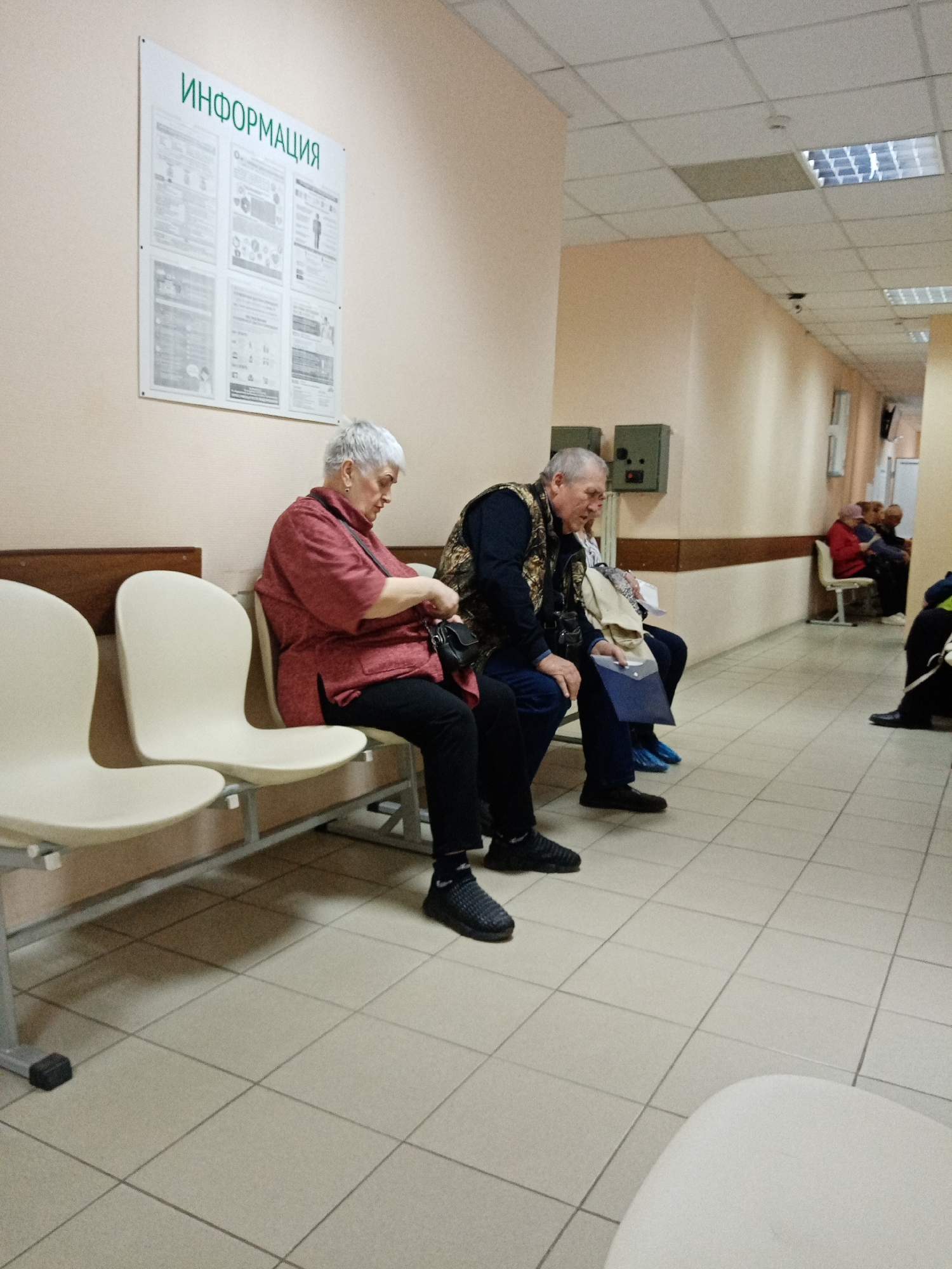 The photograph depicts a waiting area, likely in a medical clinic or hospital, in Elektrostal, Russia. The room has tiled floors, pale peach-colored walls, and fluorescent lighting. A sign with the word "ИНФОРМАЦИЯ" (Information) is posted on the wall.
Several people are waiting. A woman with short gray hair is sitting in a light-colored plastic chair wearing a red shirt and black pants. A man in a dark jacket with a patterned vest is seated beside her, holding a blue folder. Further down the hallway, more people are seated on chairs against the wall. One person near the end of the hallway is wearing red.