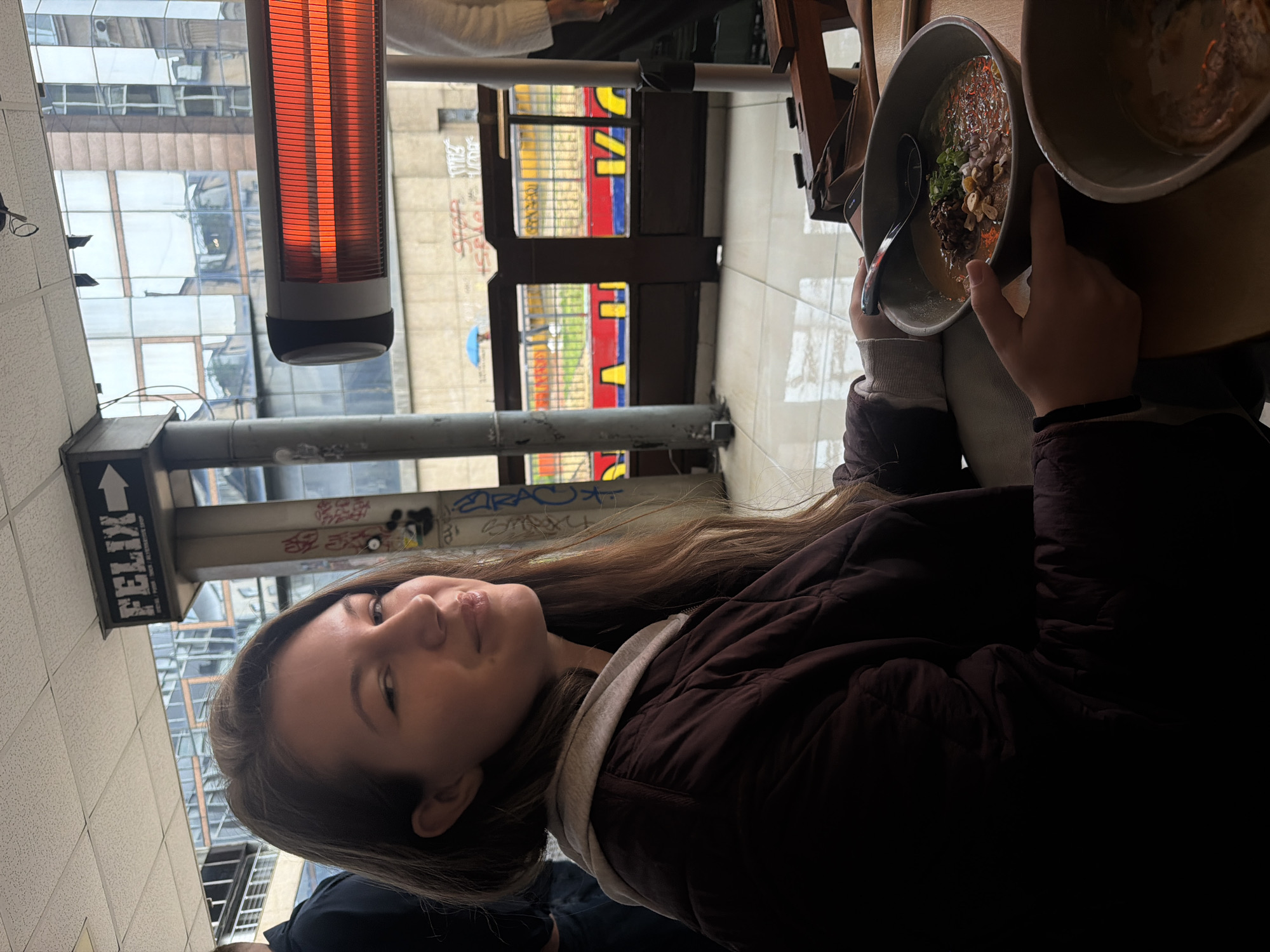A young woman in a brown jacket is seated at a table, holding a bowl of food. A second bowl sits on the table nearby. The location is an outdoor patio restaurant. Overhead, a heat lamp is mounted on a support structure. The sign "FELIX" is visible. Through an opening, city buildings are seen, indicating an urban environment. The colorful exterior wall of the Felix Bar & Kitchen, located in Belgrade, Serbia, is apparent in the background.