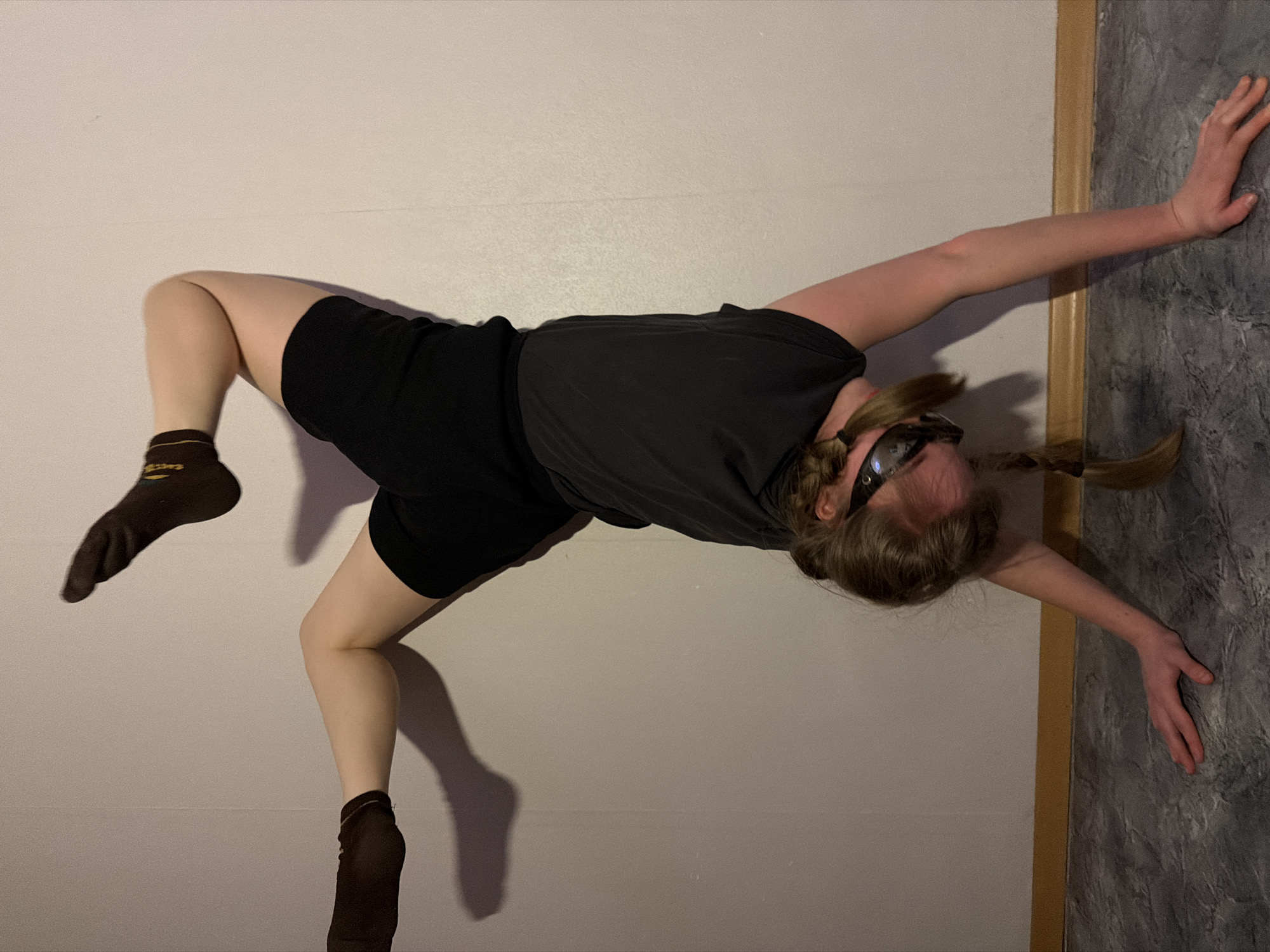 A person is positioned on a wall, appearing to hold themselves up using their arms. They are wearing a dark-colored t-shirt, black shorts, and dark socks. The person is also wearing something that looks like goggles or a headpiece. Their hair is in braids. The background consists of a light-colored wall and a dark, textured surface, possibly another wall or decorative panel, edged with a wooden trim. There is no clear indication of the city of Perm, Russia or its location in the image.