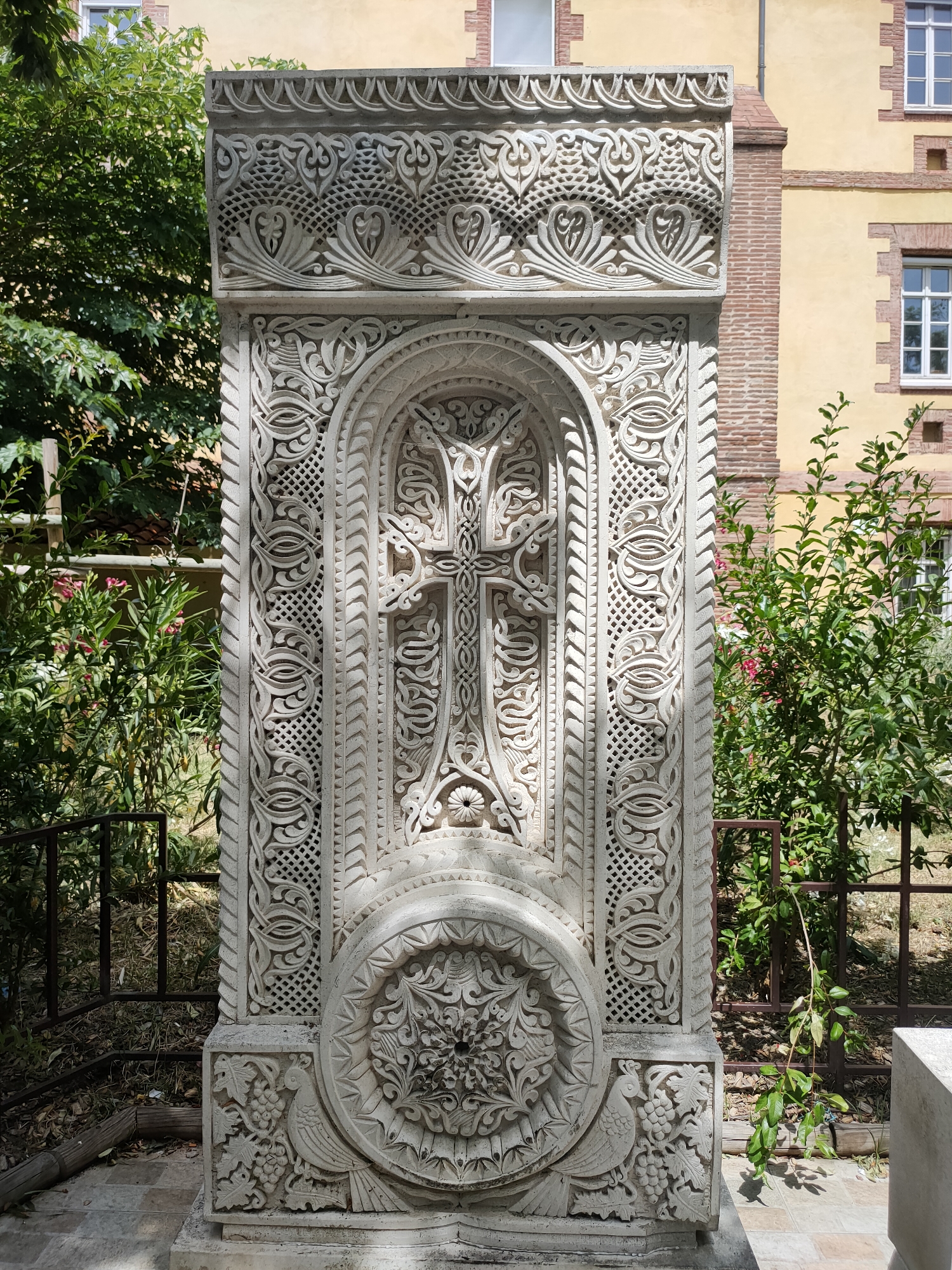 The image shows a detailed stone carving of a cross, set against a building with yellow walls and red brick accents. The carving is ornate, with intricate patterns and floral motifs. The cross itself is elaborate, with decorative elements within its structure. Below the cross, there is a circular design featuring a stylized bird and grapevines. The stone monument is positioned outdoors, with green foliage and a metal fence visible in the background. This sculpture is located at the Armenian Apostolic Church of Saint John the Baptist in Perpignan, France.