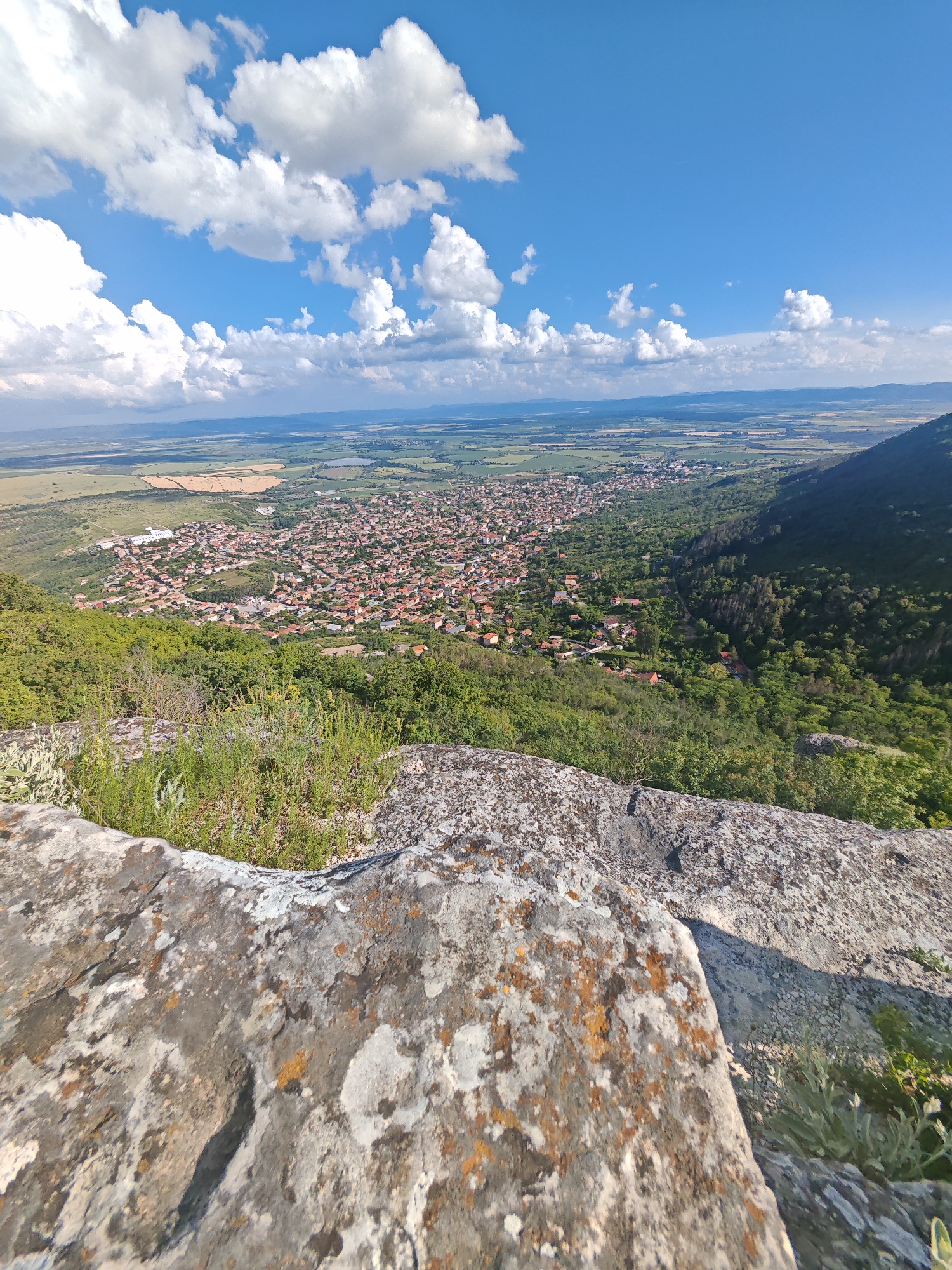 This image captures a panoramic view of the city of Shumen, Bulgaria, from a high vantage point. The upper third of the frame shows a vibrant blue sky dotted with numerous large, white cumulus clouds. In the immediate foreground, a rugged, grey rock surface, characterized by textured contours and patches of orange, white, and grey lichen, occupies the bottom quarter. Sparse green and light greyish-green vegetation grows in the crevices of the rock.
Below the foreground rock and extending into the midground, the city of Shumen unfolds, densely packed with buildings primarily featuring red-tiled roofs. Green trees and other vegetation are interspersed throughout the urban landscape. To the right of the city, a deep, heavily forested valley or gorge descends, leading to a prominent, dark green, tree-covered mountain slope on the far right. Beyond the city and valley, the background reveals a vast plain composed of various agricultural fields in shades of green, yellow, and brown, stretching towards a distant, hazy mountain range under a lighter blue horizon. The scene is brightly lit by natural daylight, casting clear shadows and highlighting surface textures. No individuals or specific actions are visible.