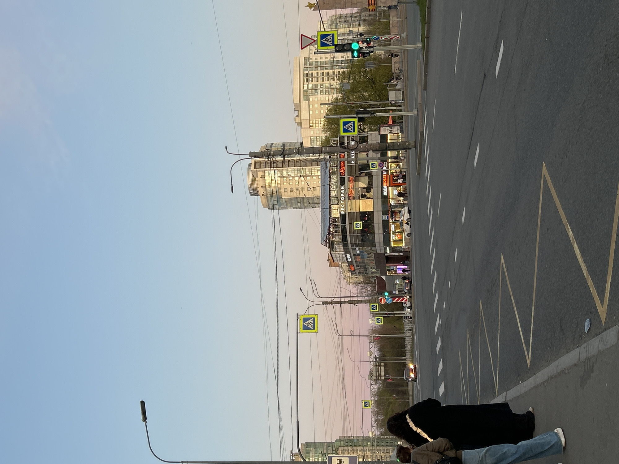 A street scene in Saint Petersburg, Russia, featuring an avenue on the right side of the frame with clear road markings and a pedestrian crossing. Various buildings line the street, with visible shops and restaurants. Streetlights and pedestrian crossing signs are present. Two individuals are lying on the sidewalk on the left side of the frame. One individual is wearing jeans and white shoes, while the other is wearing a long black coat. The overall ambiance suggests it is either late in the day or an overcast day due to the soft lighting.