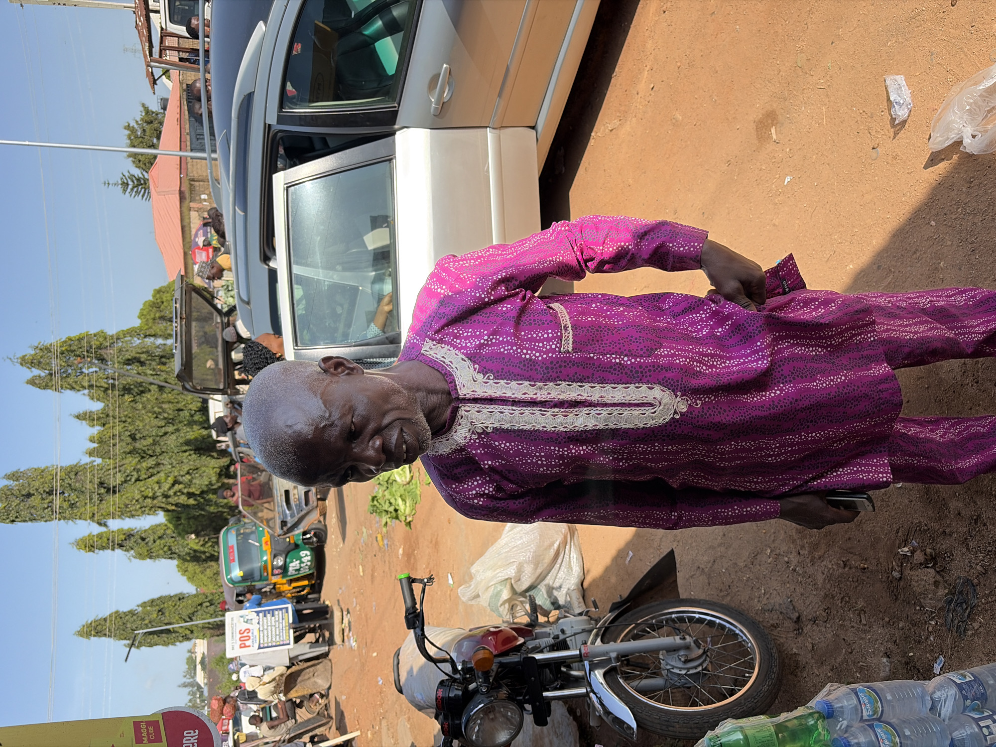 The image depicts an outdoor scene in Gura Top, Nigeria, with a man in a vibrant purple and white patterned traditional outfit standing in the foreground. He has a shaved head and appears to be looking to his left. To his left, a red and chrome motorcycle is parked, with a white sack behind it. The ground is made of red dirt. In the background, several vehicles are visible, including a silver car with its doors open, a green and yellow tricycle, and other assorted vehicles. A sign that reads "POS" is also visible in the background, as are several tall trees. The sky is clear and blue, suggesting a sunny day. There are also bottles of water stacked in the lower left corner of the image.