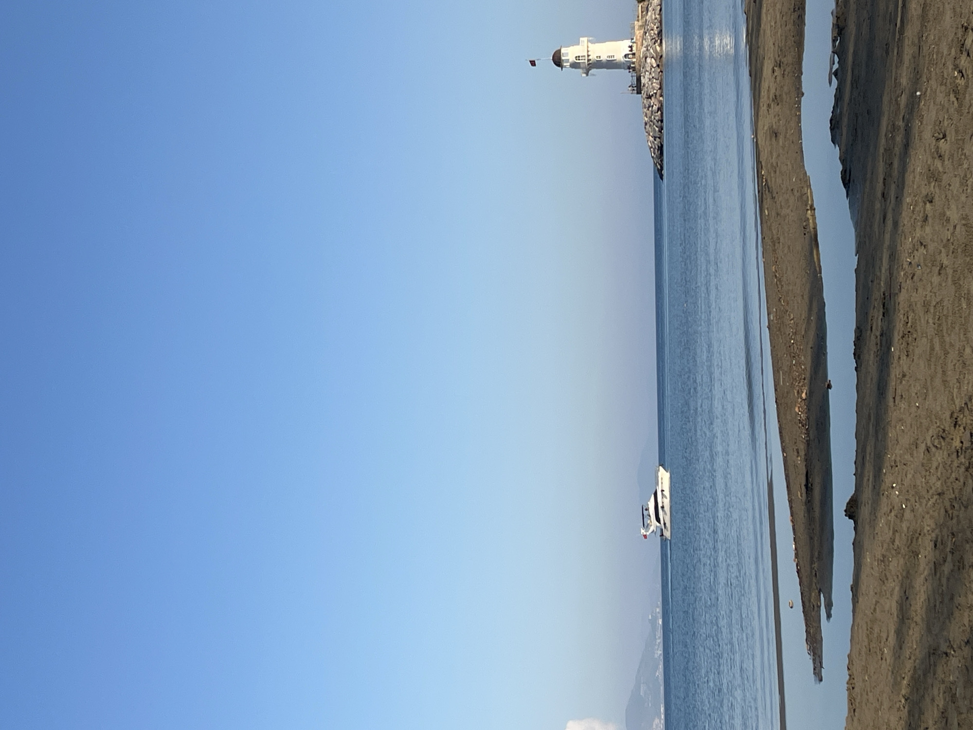 The image captures a scenic view in Alanya, Turkey. A clear, blue sky dominates the left side of the frame. The right side features the coastline, with a sandy beach transitioning into the ocean. A white lighthouse sits on a rocky outcrop at the edge of the water. A boat is sailing on the water, its white hull contrasting against the blue of the sea.