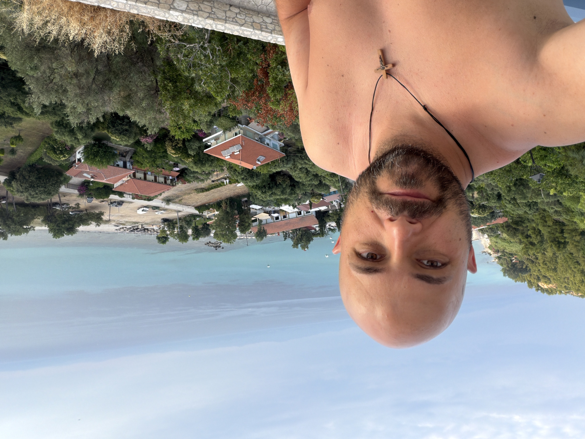 A man with a shaved head and beard takes a selfie on a balcony overlooking a coastal town in Kassandra Municipal Unit, Greece. He is wearing a necklace with a wooden cross. The man is shirtless, revealing his chest and shoulder. In the background, there are houses with red roofs, lush green trees, and the turquoise sea. The sky is partly cloudy. The scene is captured from a high vantage point, suggesting the man is on an elevated terrace or building. The image conveys a sense of relaxation and vacation in a Mediterranean setting.