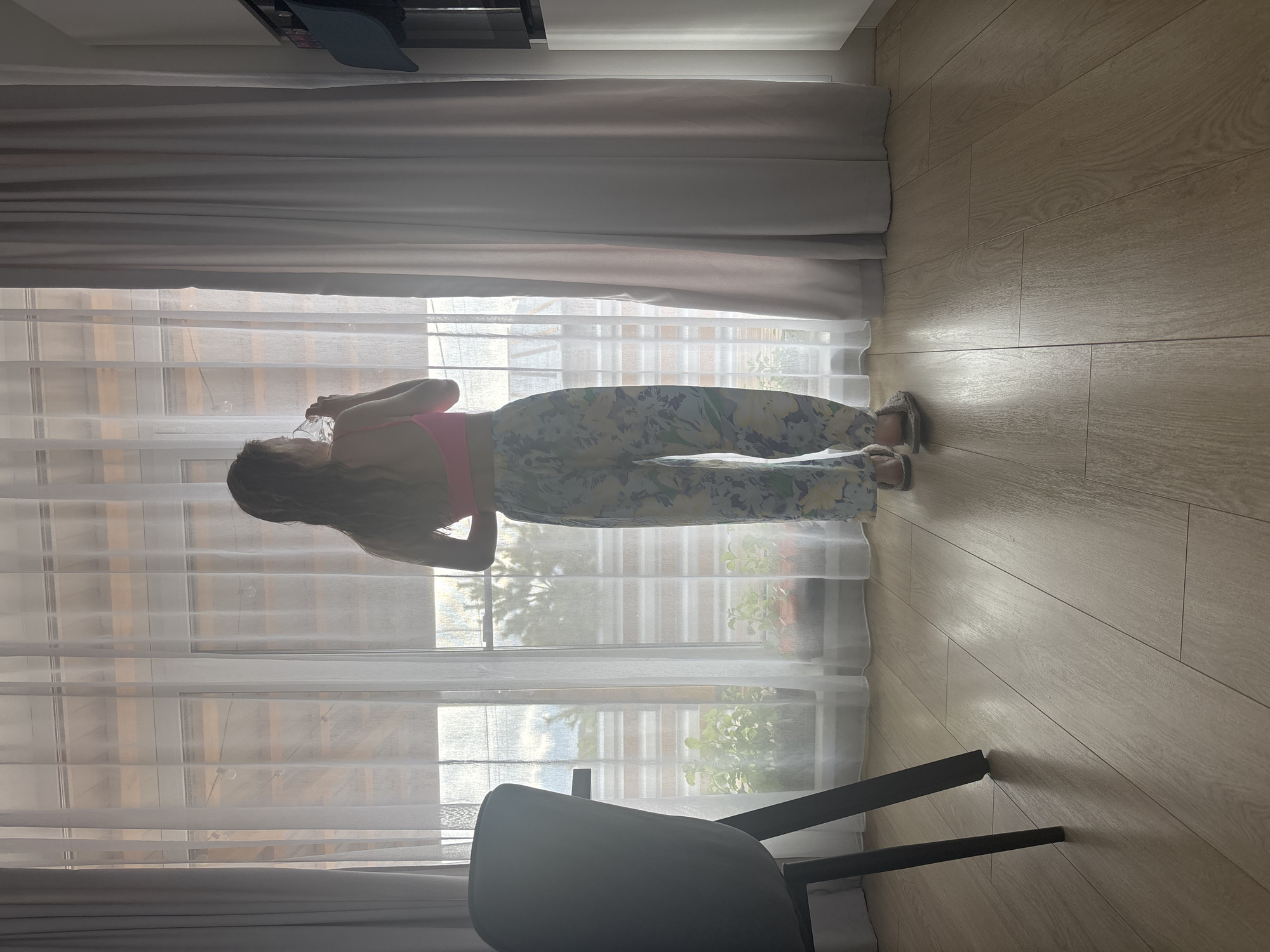 A woman stands in front of a window inside a building. She is wearing a pink top and floral-patterned pants, and she is holding a clear glass up to her face. Behind her is a white, sheer curtain covering a window. Outside the window, there are green plants and a building. The floor is light-colored wood. A dark gray chair is in the foreground. The location is the city of Chaikovsky, Russia.