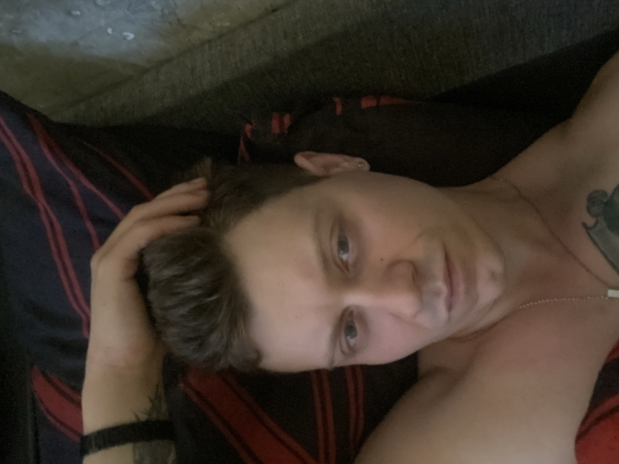 A person is lying down and looking at the camera. Their hand is on their head, and they are wearing a necklace. The background consists of a dark blanket with red stripes, and a grey textured surface, possibly a couch. The image does not contain any discernible information about the location in the city of Samara, Russia.