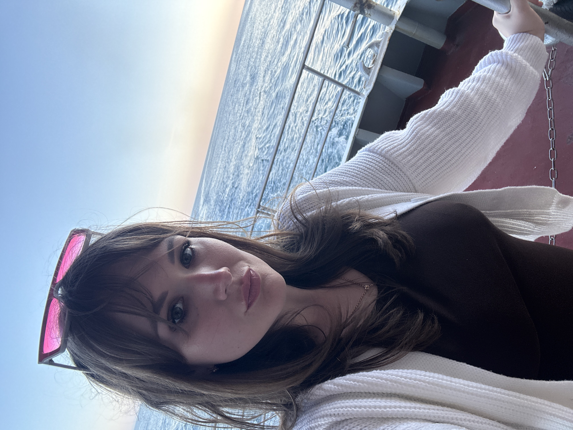 A woman is positioned on what appears to be a ship or boat, holding onto a railing. The railing and ship infrastructure are visible. In the background is the sea and a bright sky. The woman is wearing sunglasses perched on her head, a brown top, and a white knitted cardigan. A gold chain necklace is visible around her neck. The image may have been taken in Гаспринский поселковый совет, Ukraine.