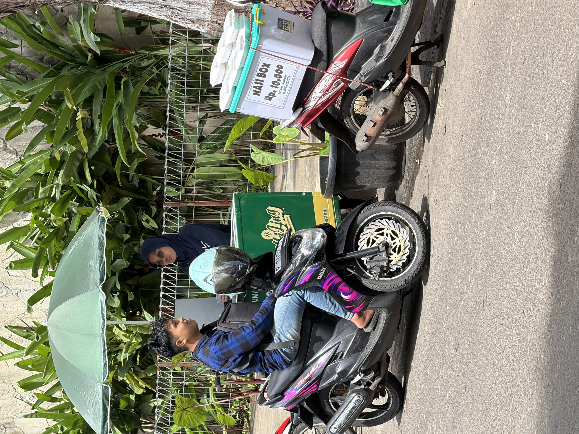 The scene is an outdoor urban environment in South Jakarta, Indonesia. A young man wearing a blue plaid shirt and jeans is sitting on a black Yamaha motorcycle. A light blue helmet rests on the front of the bike. An open, light green umbrella is positioned to the left of the motorcycle, providing shade. Behind the motorcycle, a woman in a dark hijab is partially visible. Behind them is a wire fence with foliage. Next to the Yamaha is another parked motorcycle with a white and light blue box secured to it with the label "NASI BOX Rp. 10.000". The motorcycles are parked on an asphalt road.