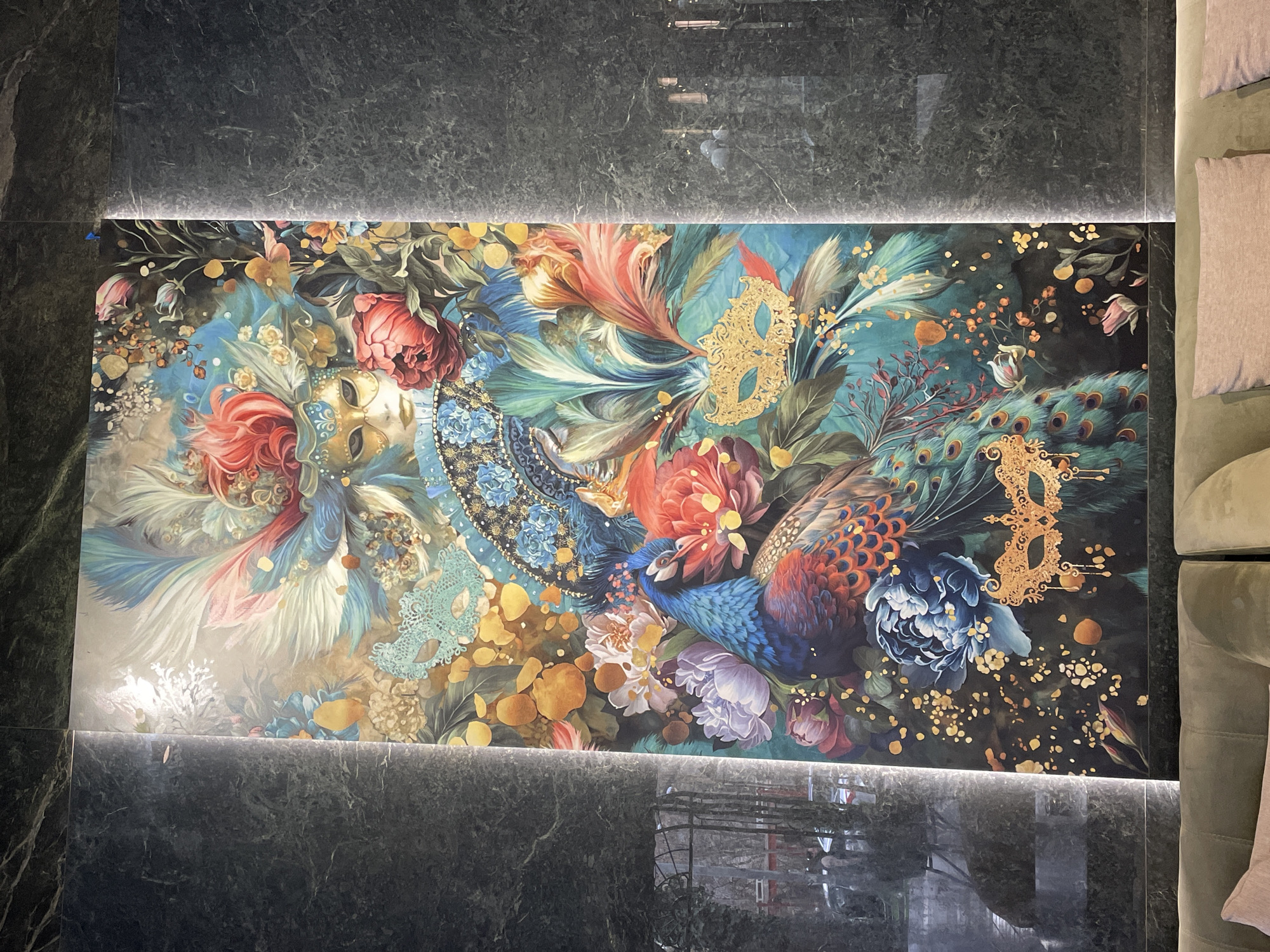 A rectangular section of a decorative tile or panel is visible, positioned on what appears to be a floor or wall surface composed of large, dark gray tiles with a speckled texture. The decorative tile features an elaborate and colorful floral design, incorporating images of peacock feathers and masquerade masks. The color palette includes blues, greens, reds, and golds. The tile is highlighted by linear LED lighting along the top and bottom edges. A portion of a green sofa with pink pillows is visible in the upper right of the frame. The location is inferred to be in Yerevan, Armenia, based on the prompt's request to identify the location.