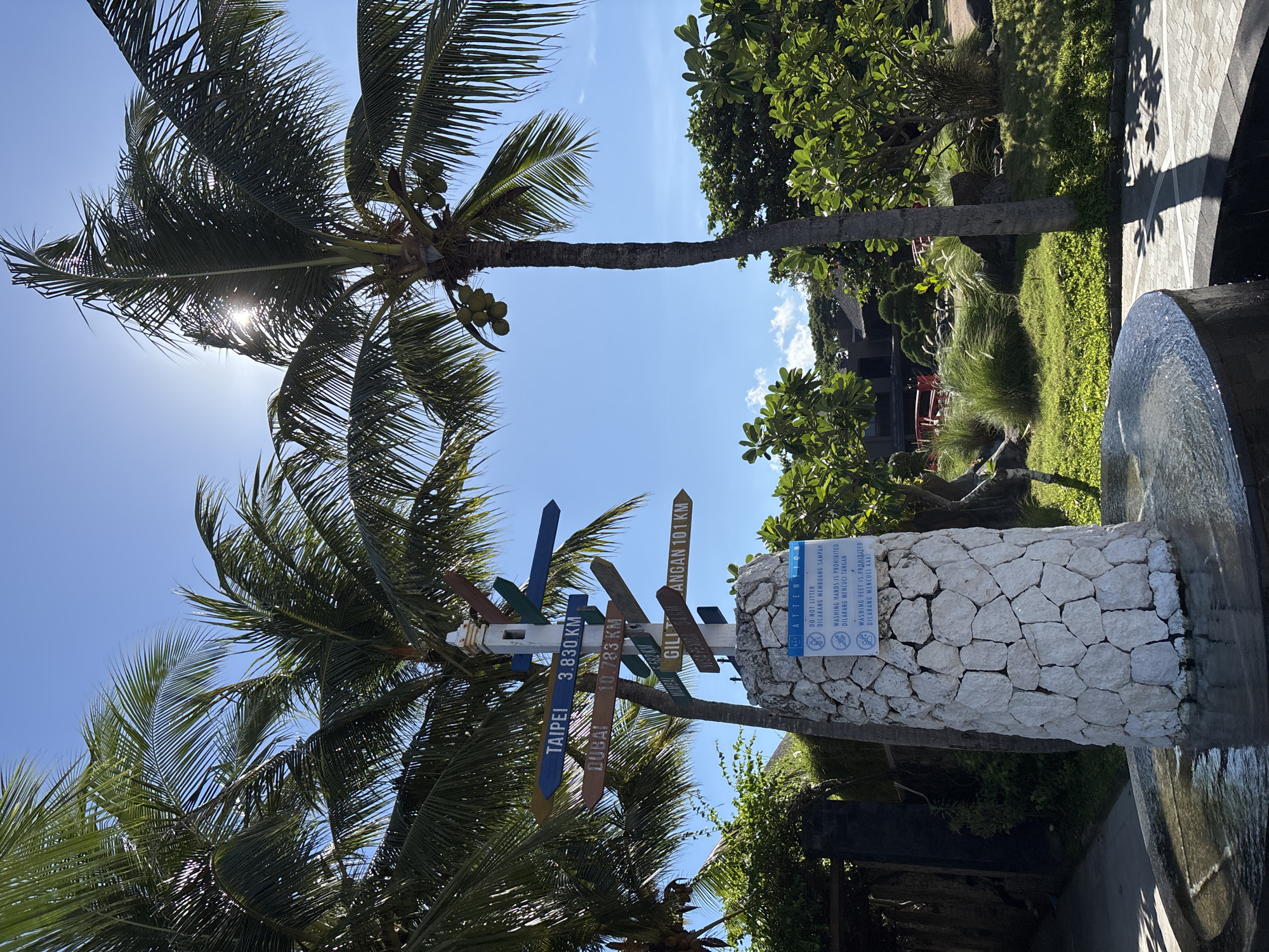 This full-shot image captures a vibrant tropical outdoor scene under a clear blue sky on a bright, sunny day. Two prominent palm trees, laden with coconuts, frame the left side of the frame, with direct sunlight creating a lens flare through their fronds.
In the midground, a wooden directional signpost displays multiple arrow-shaped signs indicating various destinations and their distances. Visible text on these signs includes "TAIPEI 3.830 KM", "DUBAI 10.783 KM", and "GILI ANGAN 101 KM". To the right of the signpost, a cylindrical pillar made of white, rough-hewn stones is visible. Affixed to this pillar is a blue and white "ATTENTION" sign.
The "ATTENTION" sign outlines rules in both English and Indonesian. The text reads: "ATTENTION", "DO NOT LITTER", "DILARANG MEMBUANG SAMPAH", "WASHING HANDS IS PROHIBITED", "DILARANG MENCUCI TANGAN", "WASHING FEET IS PROHIBITED", and "DILARANG MENCUCI KAKI". Icons depicting forbidden actions accompany the text.
The right side of the image features a paved area, lush green grass, and various tropical plants, suggesting a well-maintained garden or resort environment. A dark, rounded water feature or fountain basin is partially visible in the bottom right corner. The presence of "GILI ANGAN" on the directional sign and the Indonesian text on the attention sign strongly indicate a location in Indonesia. No people are visible in the scene.