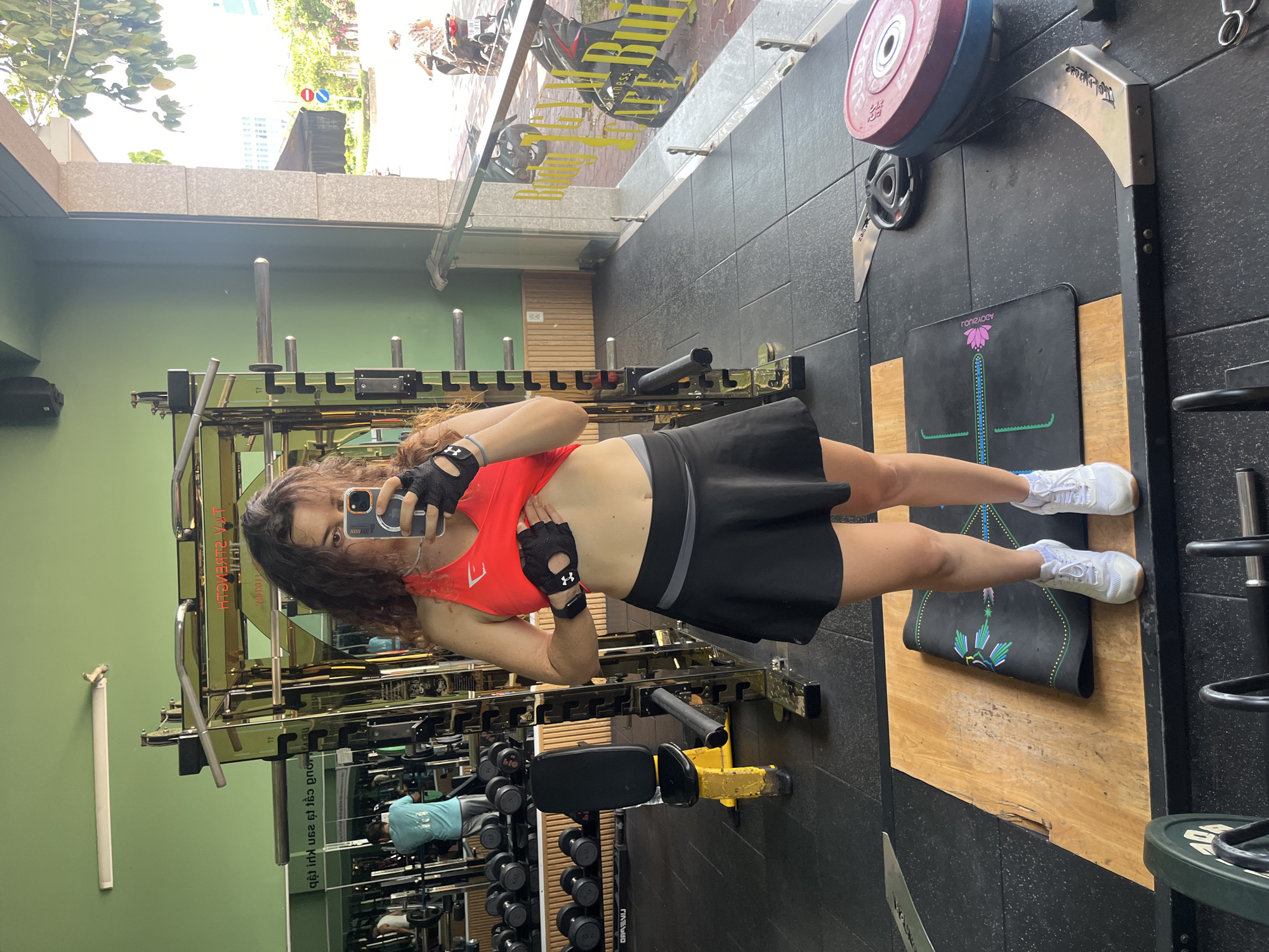 A woman with curly brown hair takes a mirror selfie in a gym located in Phan Thiết, Vietnam. She is wearing a red athletic top, black workout gloves, and a black skirt. Her midriff is exposed, revealing a toned stomach. She is holding her phone up to take the photo. In the background, gym equipment such as weight racks, benches, and dumbbells are visible. Through a glass window on the right side of the image, a street scene in Phan Thiết can be observed, with a person riding a motorbike and trees. The gym appears to be well-lit and equipped for a variety of exercises.