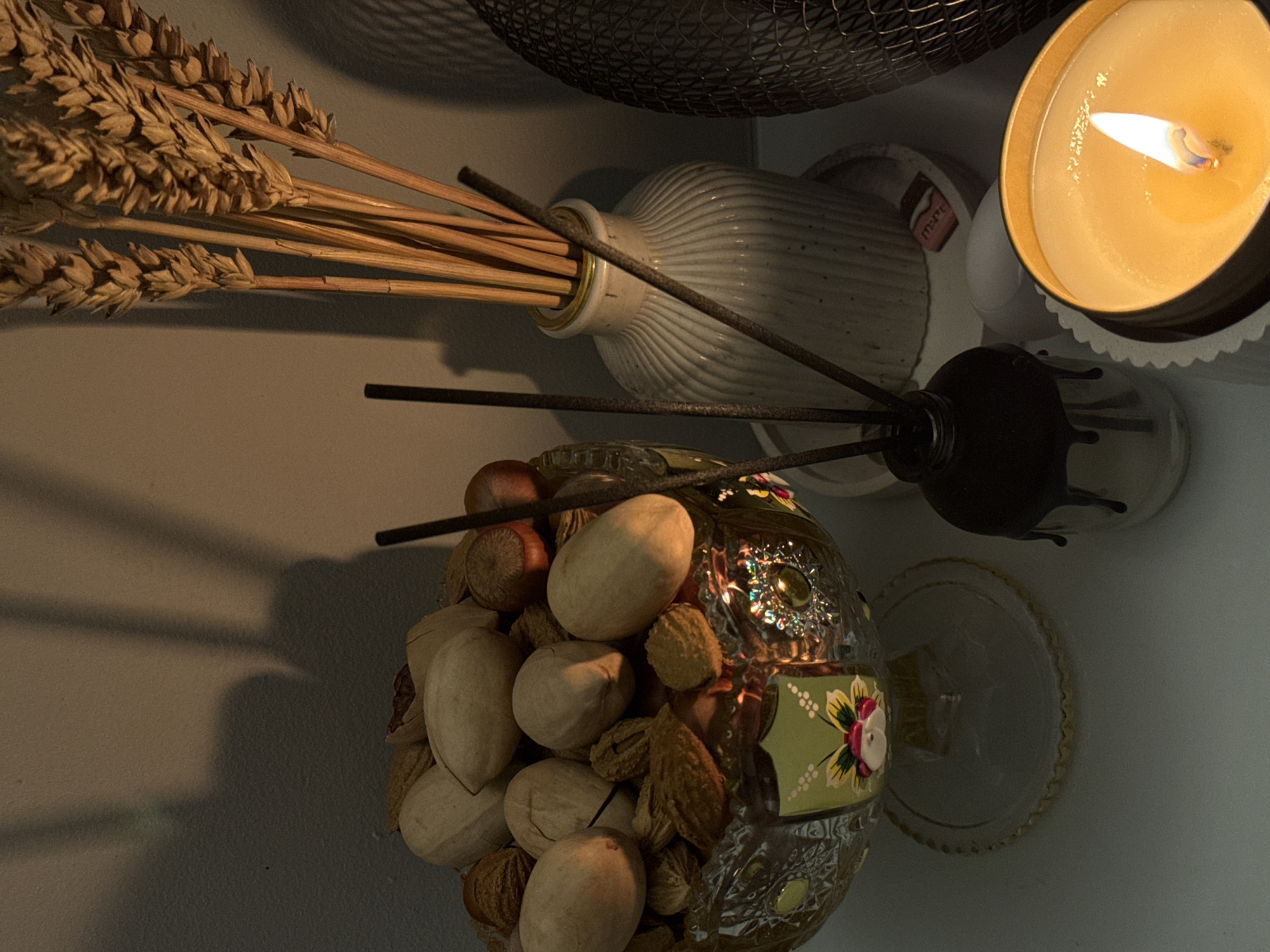 The image shows a still life composition of various objects. On the upper left, there is a bundle of wheat stalks placed in a light-colored vase with ribbed texture. Above the vase is a dark, wire-like object that is out of focus. To the right of the vase, there is a lit candle in a tin container, with visible melted wax around the wick. Below and to the right of the vase is a clear glass diffuser bottle with dark reeds inserted into it. Beneath the diffuser is a clear glass ornamental bowl filled with nuts and other dry botanicals. The background is a light gray wall with subtle shading.