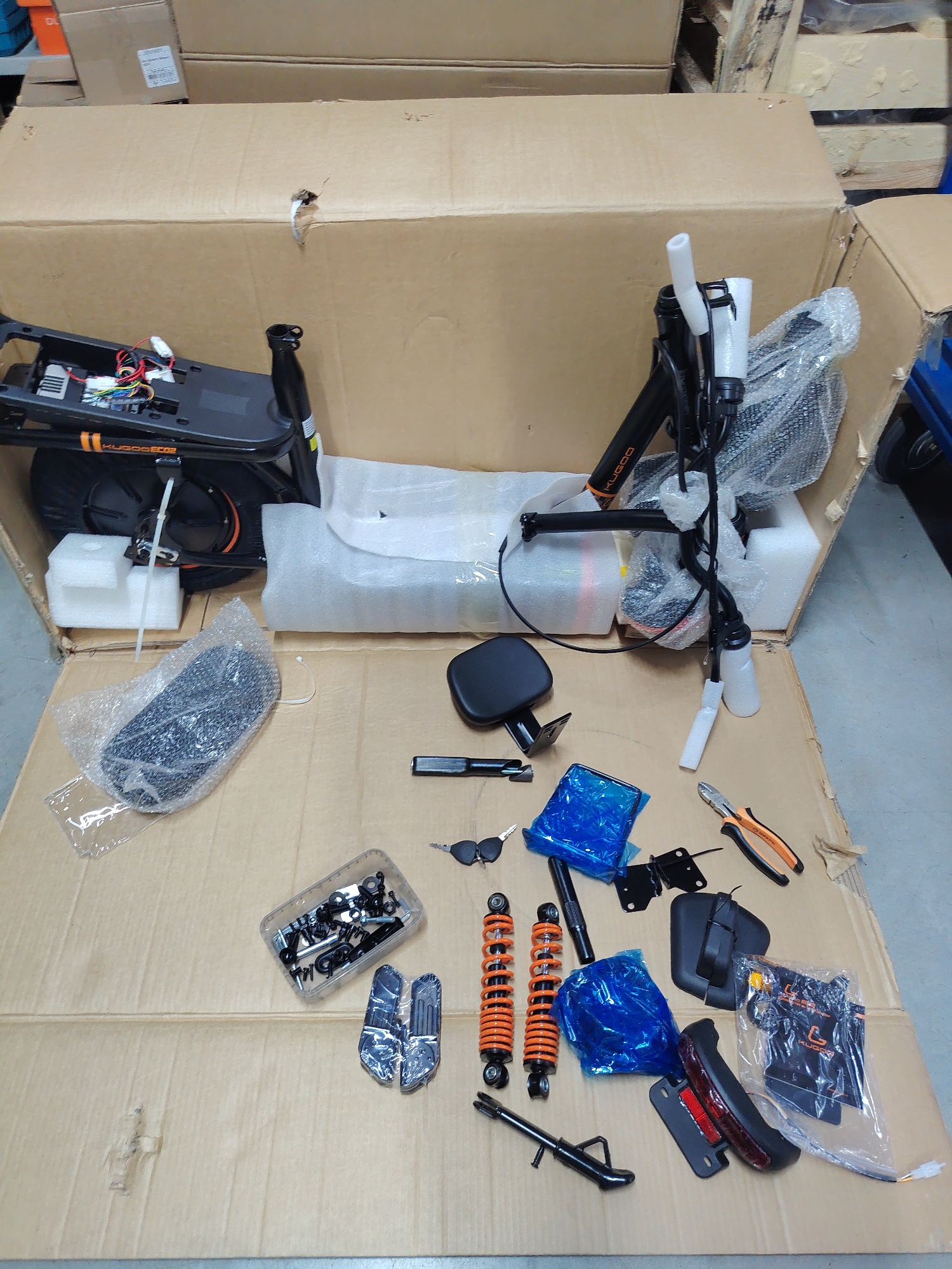 The image shows the contents of a large cardboard box, seemingly a disassembled electric scooter, laid out on a cardboard surface. The main body of the scooter, including the deck with electronics and wiring, and the handlebars with part of the steering column, are prominently displayed. Various parts are scattered around: shock absorbers, a seat, keys, a small bag of hardware, a tool (pliers), a rear light assembly, and other smaller components. The environment appears to be a storage or warehouse setting, suggested by the presence of other cardboard boxes and pallets in the background. No people or specific locations in Kazan, Russia are visible or implied in the image.