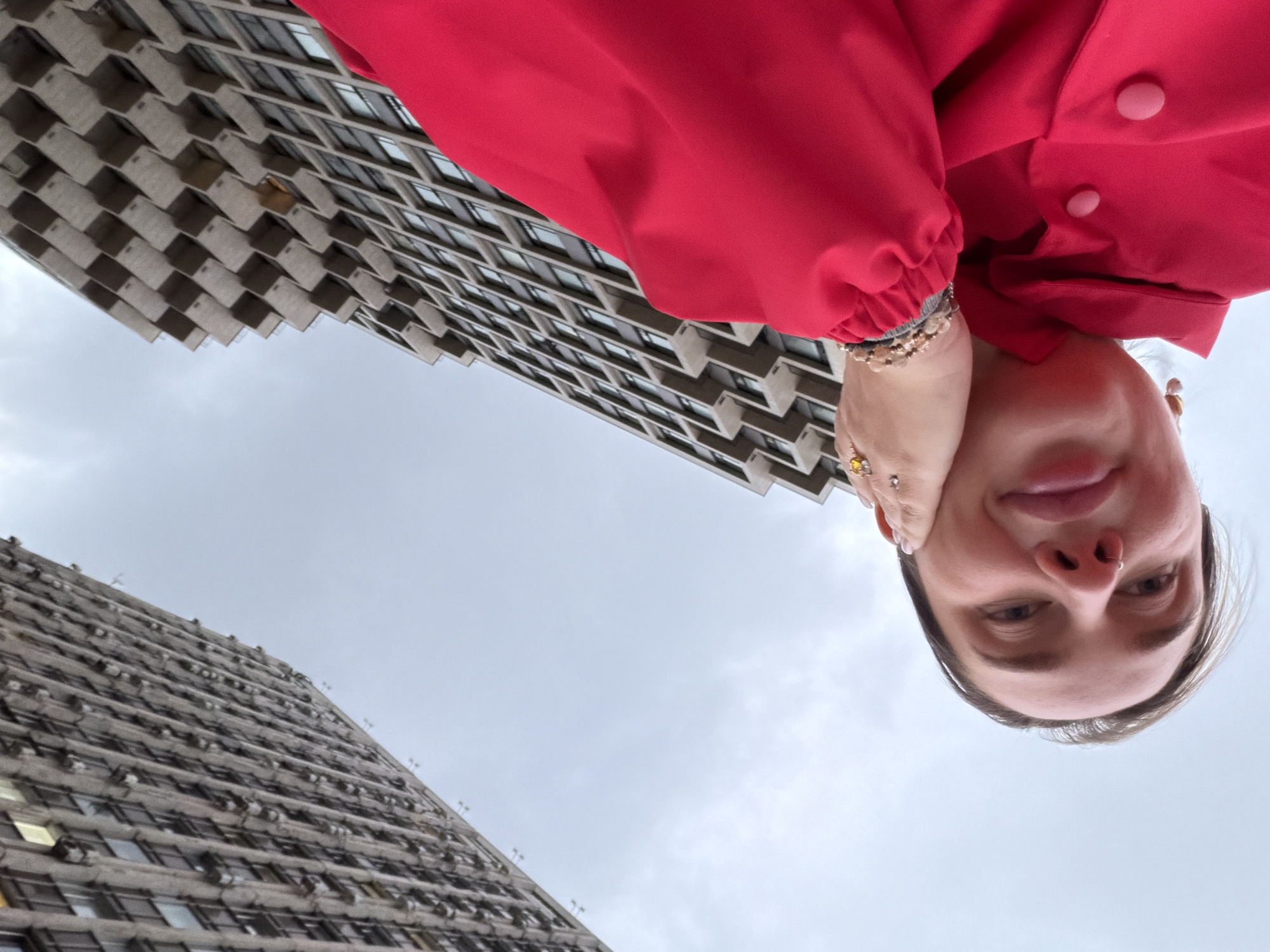 The image displays a low-angle, rotated perspective featuring a person and two tall buildings against an overcast sky in Moscow, Russia. The entire photograph is rotated 90 degrees clockwise, resulting in the buildings appearing to lean horizontally and the person's face being oriented upside down.
The primary subject is a woman, occupying the central-right portion of the frame. She looks towards the camera with a neutral to slightly smiling expression, her face appearing upside down relative to the ground. She wears a vibrant red jacket or coat, which covers the upper right corner, and her right hand, adorned with multiple bracelets and rings, rests near her chin. A septum piercing is visible on her nose.
In the background, two imposing high-rise buildings stand against a cloudy, gray sky. The closer building, on the upper left, exhibits a distinctive Brutalist or modernist architectural style, characterized by a complex, repeating pattern of concrete projections and recessed windows, creating a textured, almost waffle-like facade. The second building, further in the distance on the lower left, shares a similar architectural language, featuring rows of windows and horizontal concrete elements.
The scene depicts an urban setting under an overcast sky, suggesting a daytime shot without direct sunlight. The photo appears to be a selfie taken from a low vantage point, looking upwards towards the towering structures. No visible text is present in the image.