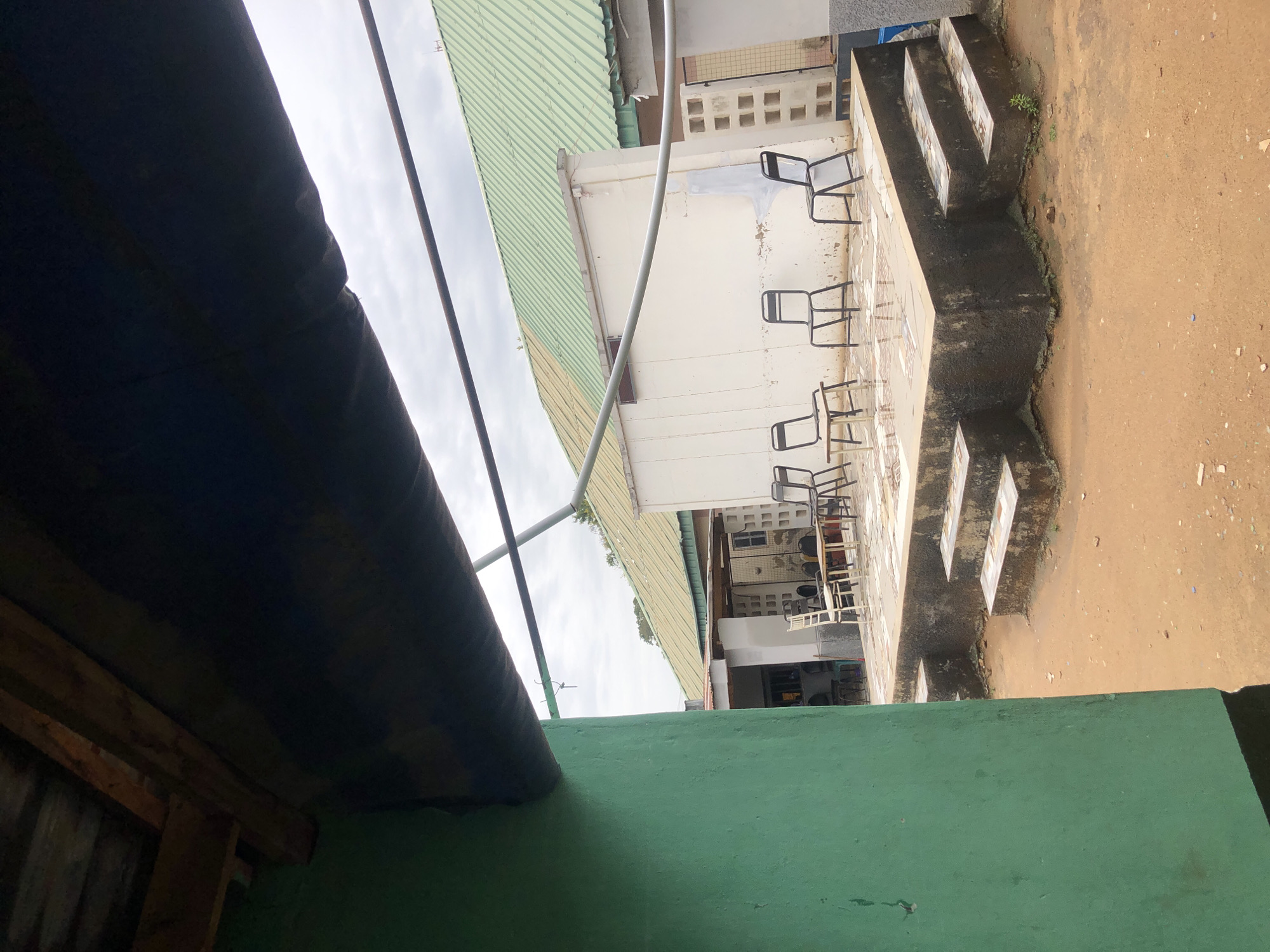 The image presents an outdoor scene, likely a courtyard or patio, viewed from a slightly elevated vantage point or from beneath an overhang. In the immediate foreground on the left, a dark corrugated roof or awning casts a shadow, with a distinct green wall or railing visible below it, along the bottom of the frame. A thin metal pole or pipe extends diagonally from the upper left across the upper middle portion of the image.
The central focus of the scene is a concrete or tiled patio area furnished with multiple chairs. These chairs are predominantly metal-framed, mostly black, with some featuring lighter-colored seats. A small table is also visible among a cluster of chairs. Behind this seating area stand white buildings topped with light green corrugated metal roofs. One building face displays a pattern of square vents or openings.
To the right of the patio, a set of concrete steps descends to an uneven, reddish-brown dirt ground area that occupies the right side of the image. This ground appears somewhat barren, with a few small patches of green vegetation and scattered debris.
The sky, visible above the buildings and roofs, is overcast, indicating a cloudy daytime setting. No people are present, and therefore, no specific activity or event is depicted. The architecture, materials, and ground cover suggest a utilitarian outdoor space, possibly a waiting area, schoolyard, or communal area, potentially located in a warm or tropical climate. There is no visible text within the image.