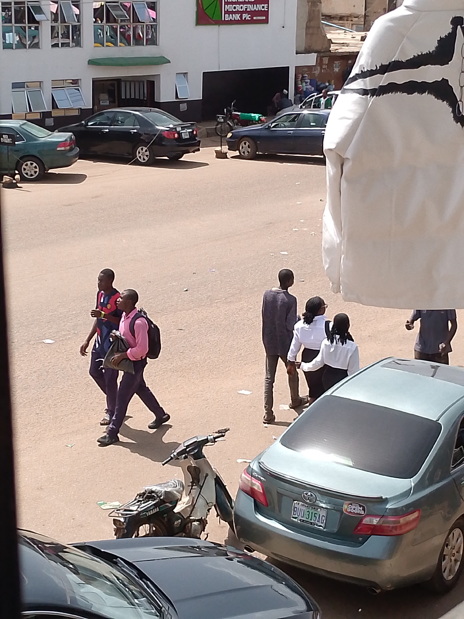 The image depicts an outdoor scene in what appears to be a commercial area in Bukuru, Nigeria. The foreground is dominated by the rear of a light blue sedan, the license plate of which reads "BUU 315AG". Two stickers are visible on the trunk of this car: one circular sticker with the words "OPEN DOORS" and another rectangular one. To the left of this car, the front of a dark-colored car is visible.
Further back, several people are walking on a paved surface. Two young men in casual attire are walking towards the right of the frame. One is wearing a blue and red shirt and dark pants, while the other, slightly ahead, is wearing a pink collared shirt, dark pants, and carrying a backpack. Behind them, a man in a grey shirt and dark pants is walking with two women, both wearing white tops and dark bottoms. Their body language suggests they are engaged in conversation.
In the background, a white building with multiple windows is visible. A sign above an entrance reads "HIGHLAND MICROFINANCE BANK PLC". Several cars are parked or driving past the building. A motorcycle is also visible parked to the left. The lighting suggests it is daytime, likely midday given the bright sunlight and shadows. The overall scene conveys a sense of everyday activity on a street in a Nigerian town.