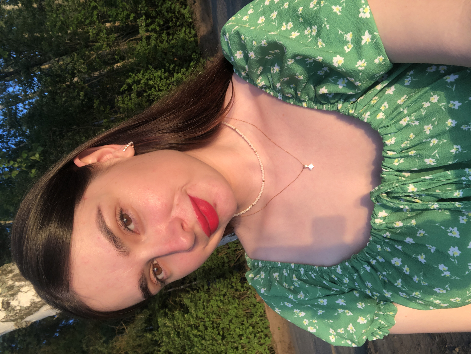 A young woman with long, dark hair and fair skin is wearing a green floral print top and posing for a picture. She is wearing red lipstick and has a small earring in her left ear. She is wearing two necklaces, one of pearls and another of gold with a small pendant. The background appears to be a green, leafy area, possibly trees. The image does not depict the location in the city of Chaikovsky, Russia, so I cannot verify the location.