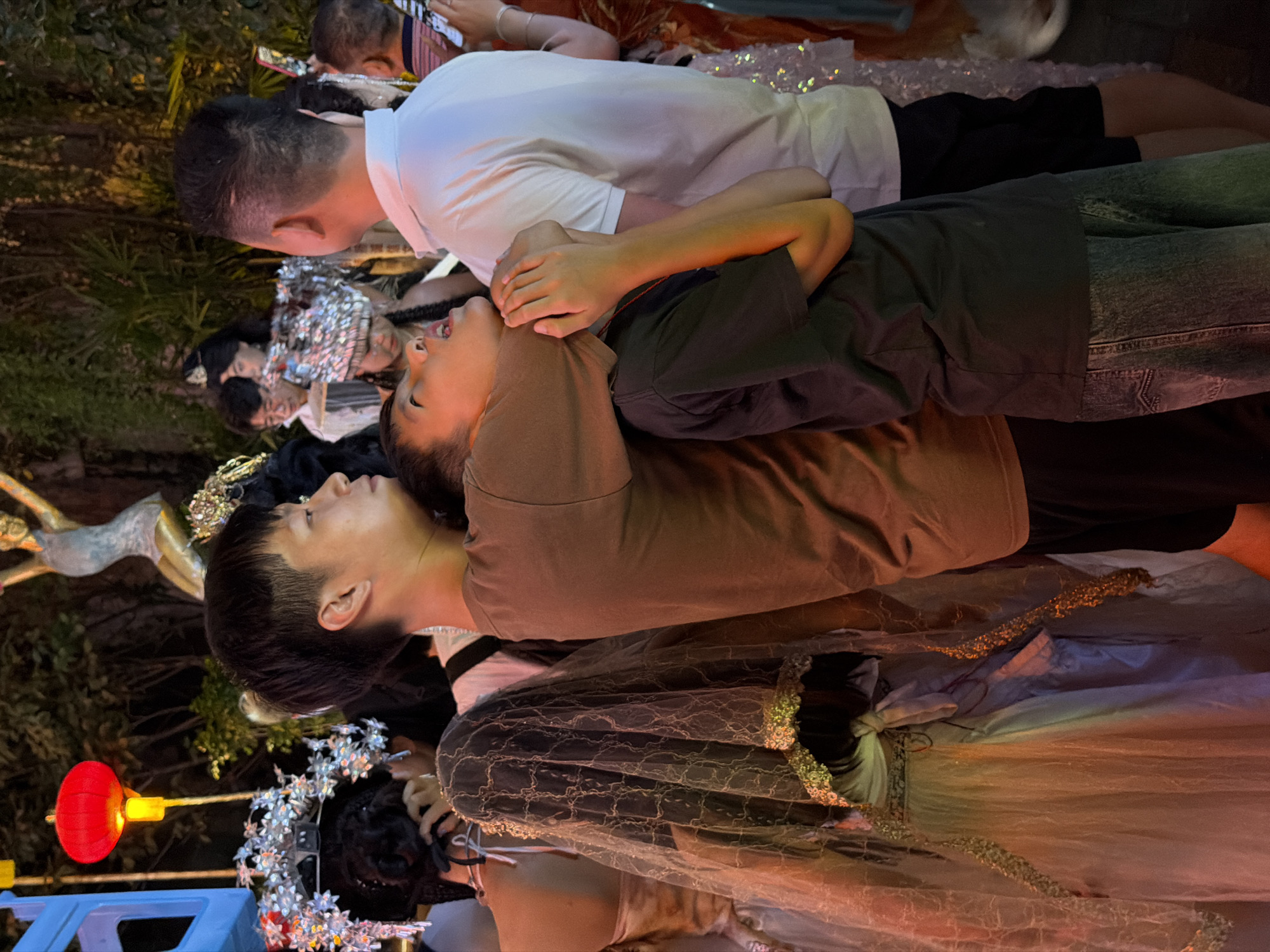 In Yuzhong District, China, a group of people are gathered at night, illuminated by a red lantern and ambient lighting. In the foreground, a young boy is embraced from behind by an older boy. The younger boy's head is tilted back, his mouth agape, and his eyes are closed. His arms are raised, with his hands clasped around the older boy's neck. The older boy is wearing a brown t-shirt and has his arms around the younger boy. To the right, a man in a white polo shirt stands with his back to the camera. Further to the left, a woman is partially visible, wearing a white dress and a decorative veil that covers her head and shoulders. Other individuals are also present in the background, some wearing elaborate headdresses. The environment appears to be outdoors, with foliage and decorative elements suggesting a festive or celebratory occasion.