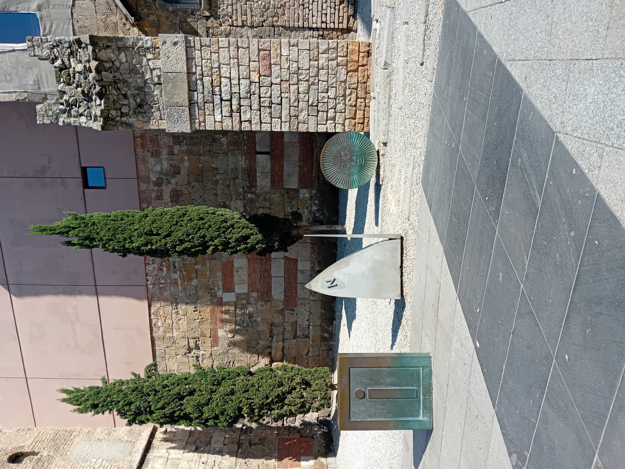 This outdoor image, captured in Barcelona, Spain, under bright daylight with clear skies, depicts a public space alongside historical and modern architecture. The strong sun casts sharp shadows from the various elements.
In the foreground, a paved walkway shows a clear division between lighter and darker grey sections. Three distinct metal objects are embedded in the lighter paving. Closest to the viewer on the right is a rectangular, verdigris-patinated metal plate featuring a central rectangular slot and a small circular detail above it. To its left, a metallic, blade-like sculpture with the number "17" engraved on its surface rises from the ground. Further back and slightly to the right, a large, circular, verdigris-colored metal disc is flush with the pavement. This disc displays a stylized design, possibly a rooster or a crest, encircled by radial lines, and faint lettering that may include "SSD".
Along the left side of the scene, a tall, slender evergreen tree, likely a cypress, stands prominently. Behind it and stretching across the mid-ground are ancient-looking stone walls, constructed from various types and sizes of stone, interspersed with sections of reddish brickwork. Green shrubbery grows at the base of these walls. To the right of the stone walls, a modern, light-colored building with a small square window is partially visible. The upper left corner reveals more rough stone structures, possibly ruins.
The scene appears to be a well-maintained public area, possibly incorporating art installations or historical markers. No people are present.