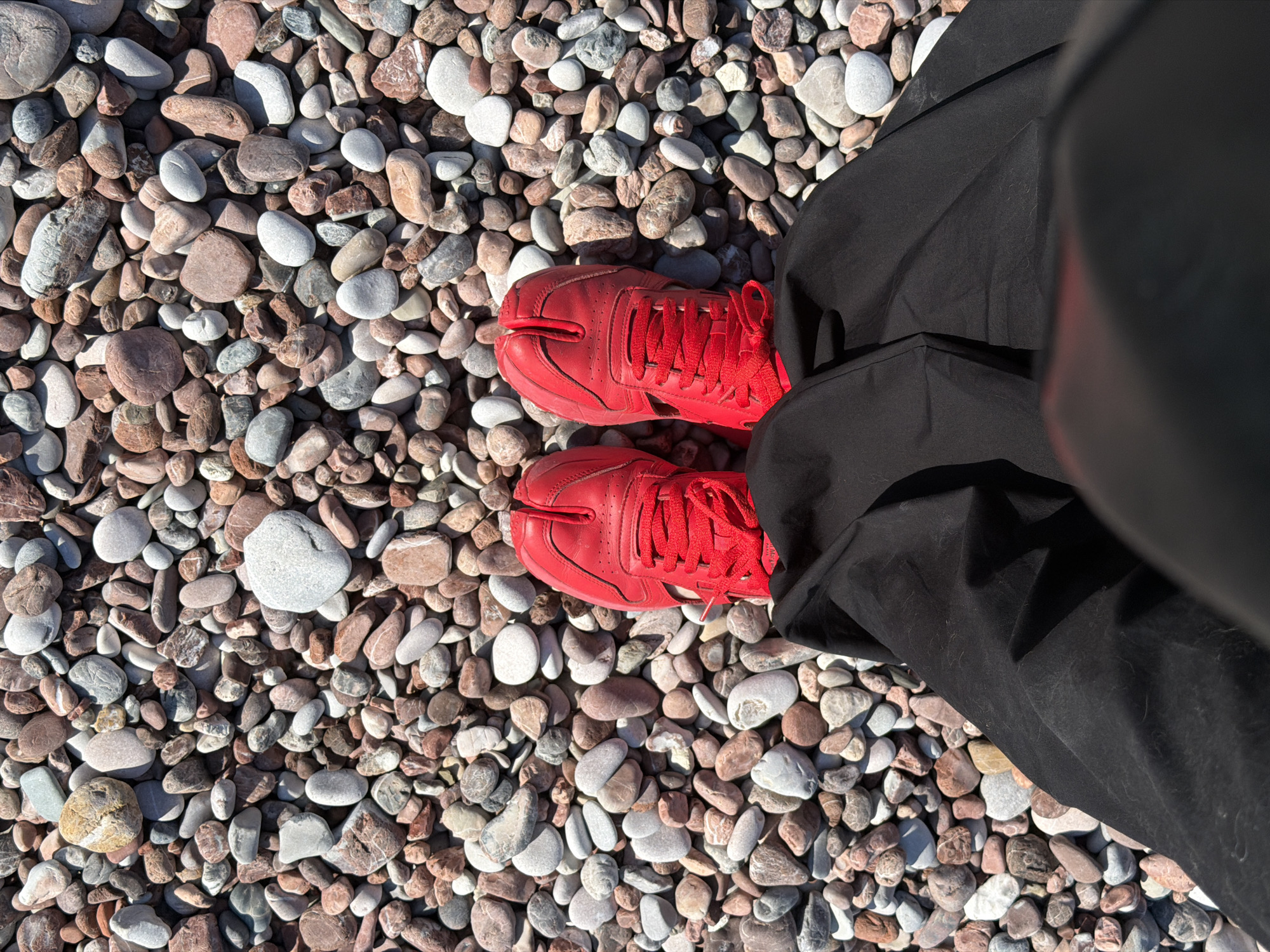 The image shows a person standing on a pebble beach in Sveti Stefan, Montenegro. The ground is covered with light-colored pebbles. The person is wearing a pair of bright red, split-toe sneakers and black pants. The perspective is looking down on the person's feet.