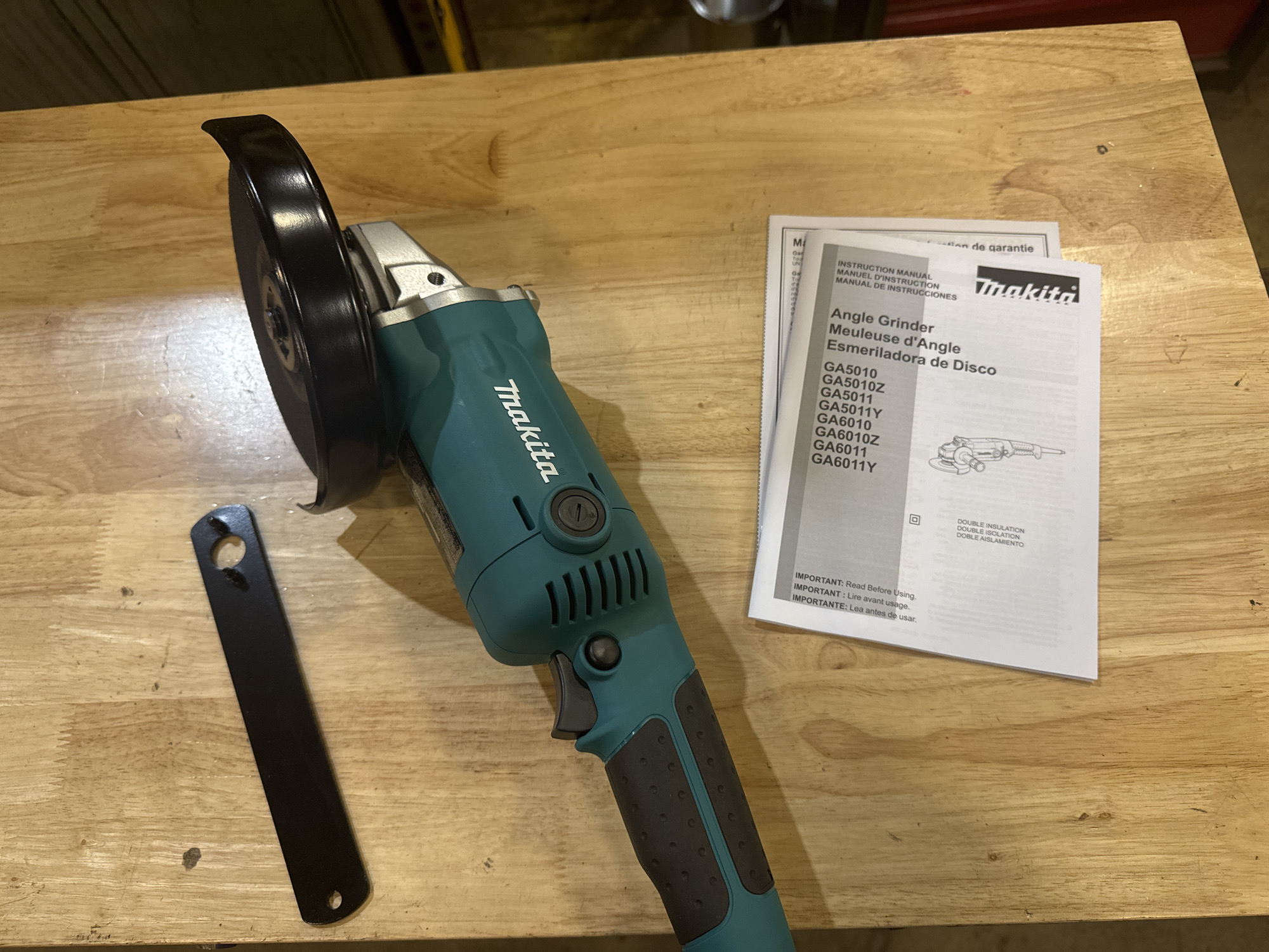 An overhead shot shows a teal and silver Makita angle grinder, a black metal wrench, and an instruction manual on a light-colored wooden surface. The grinder, positioned on the right, has a black disc guard and a black grinding disc. The wrench is on the left, elongated and black. The instruction manual is in the lower right corner, partially unfolded, displaying text and a diagram of the tool. The wooden surface appears to be a tabletop or workbench, with visible wood grain and some minor imperfections. No people or other objects are present. The scene is lit from above, casting soft shadows. The overall impression is that of a tool and its accompanying documentation laid out for use or inspection. The context is domestic or workshop. There is no information within the image that can place it in Calgary, Canada.