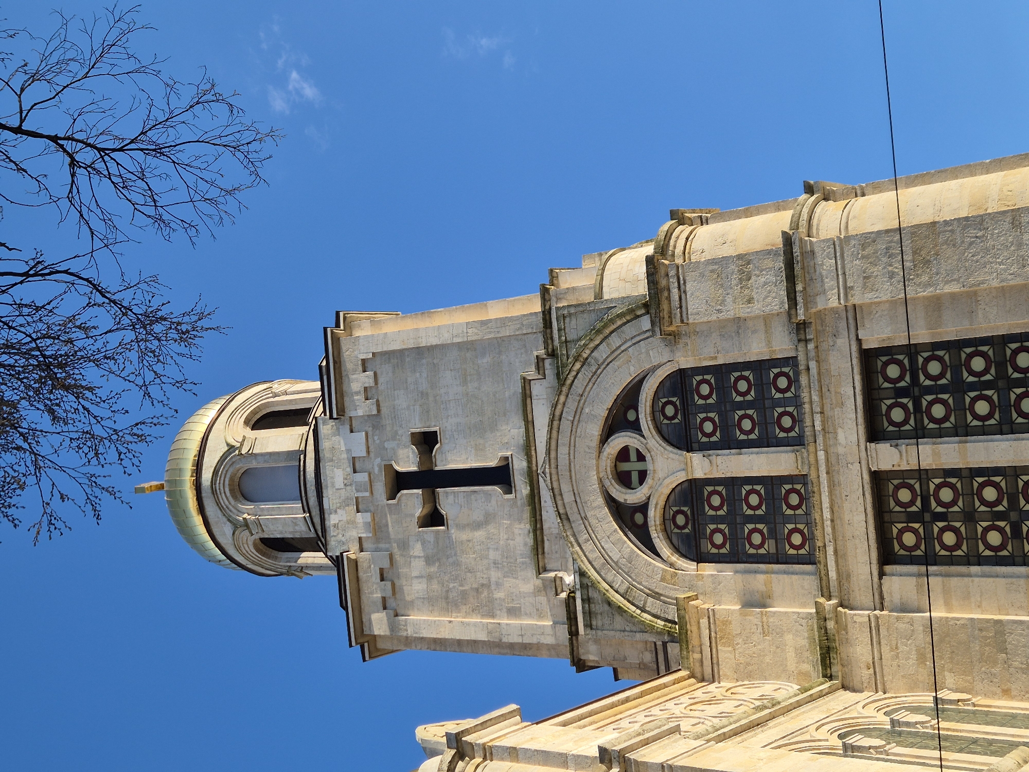 A low-angle shot captures a section of the Cathedral of the Assumption of the Virgin in Varna, Bulgaria. The cream-colored stone structure is set against a clear blue sky. Bare tree branches extend into the top left corner of the frame. The architecture is dominated by a tower featuring a dark cross, a circular window, and rows of windows with circular designs. A rounded dome with a golden tip crowns the structure. A thin, dark wire extends from the top right of the building toward the top of the frame. The image is oriented with the cathedral pointing upwards, giving a slightly tilted perspective.