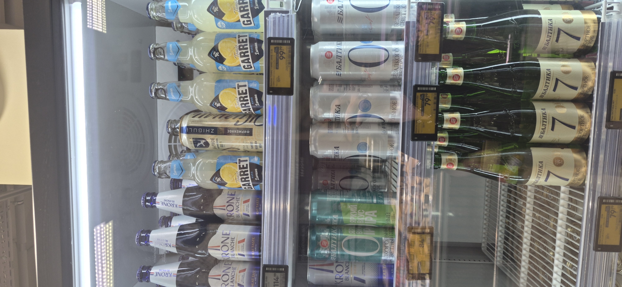 This media file captures a close-up view inside a refrigerated display case, likely at a grocery store or convenience store in Saint Petersburg, Russia. The scene is dominated by rows of beverages, primarily bottles and cans of beer and other drinks.
On the left side of the display, several glass bottles with blue and yellow labels are visible. These bottles appear to be some kind of lemon-flavored beverage, with "GARRET Lemon" prominently displayed on their labels. Below these, there are bottles with "KRONE BLANCHE" branding.
In the center of the display, there are numerous silver and green cans. The silver cans have "БАЛТИКА" written on them, with a large "0" or "O" symbol. The green cans also feature Cyrillic text, including "ЯРКИЙ ВКУС" and "ПИВА."
To the right, green glass bottles with cream and gold labels are stacked. These labels clearly show the number "7" and the text "БАЛТИКА." Some of these bottles have small red and gold circular stickers on their necks.
Several yellow price tags are visible within the display. One tag shows "99.99," another "79.99," and a third clearly reads "114.99." The presence of Cyrillic text on the products and price tags, along with the mention of Saint Petersburg, Russia, provides strong location context. The lighting suggests the store is well-lit, and the image was likely taken during the day. There are no people visible in the frame. The overall impression is a well-stocked beverage cooler ready for customers.