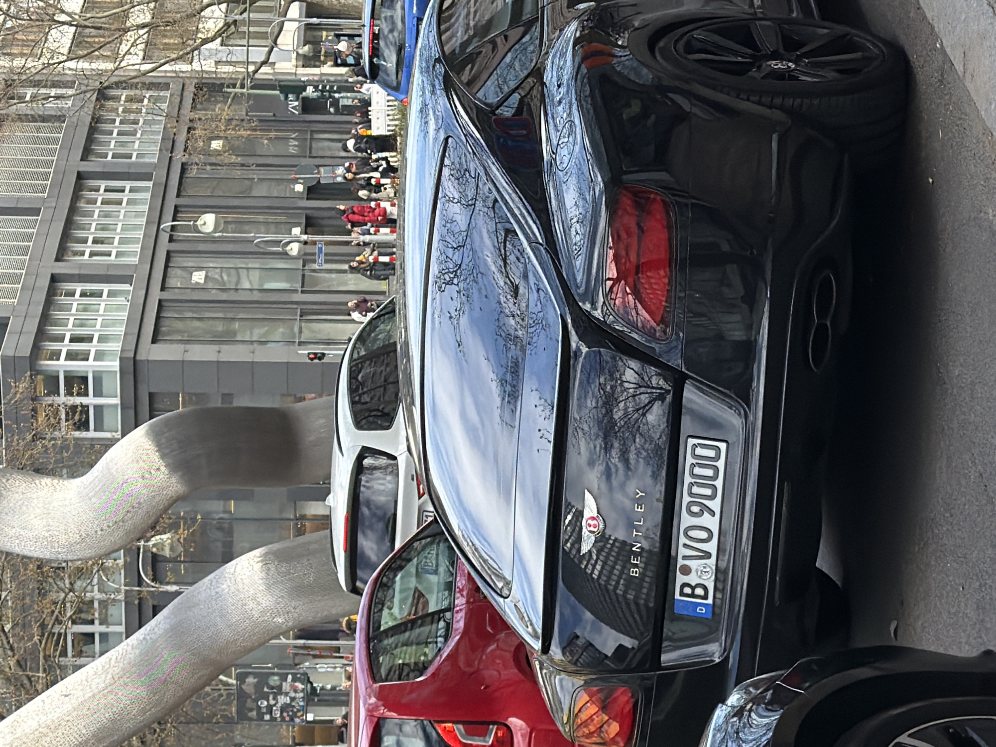 The image shows the rear of a black Bentley automobile with the license plate "B VO 9000". The car is parked on a street in Berlin, Germany. A modern art sculpture resembling intertwined tubes is visible in the background, along with a multistory building. There are people walking on the sidewalk in the distance. In the foreground, other vehicles, including a red car and another black car, are partially visible. The street is asphalt, and the sky is reflected on the surface of the car. The overall scene appears to be an urban setting during daylight.