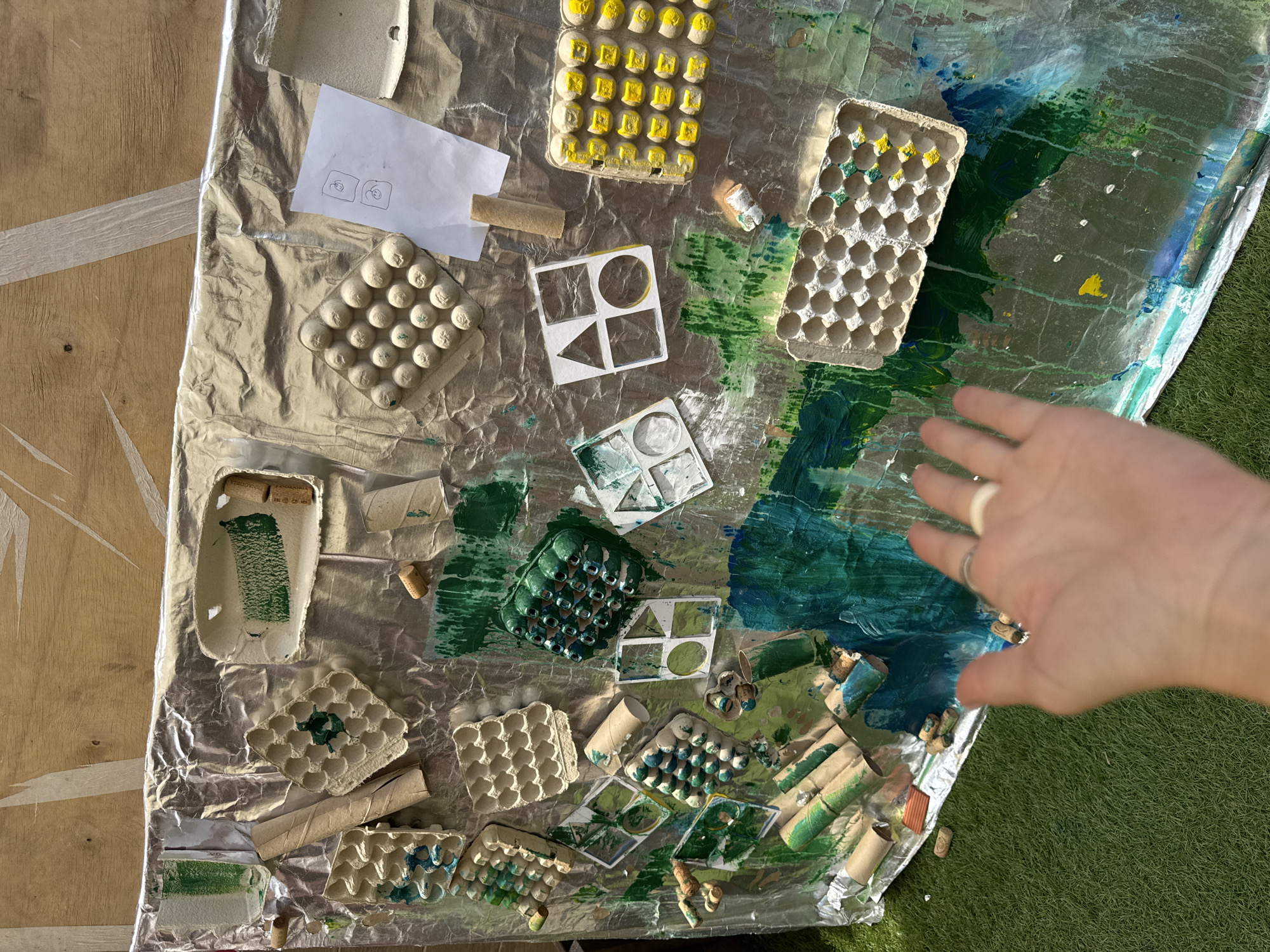 In Krasnaya Polyana, Russia, an overhead shot captures an art project display. The display is mounted on a wall with a wooden texture, covered by a large sheet of crinkled aluminum foil. The foil serves as a backdrop for various art materials, primarily egg cartons, cardboard tubes, and what appear to be painted corks. These items are arranged and adhered to the foil, with splashes and streaks of green and blue paint evident across the surface, suggesting an active creative process.
In the foreground, a person's right hand is reaching towards the artwork. The hand has rings on two fingers. The artwork itself is spread out on a grassy surface. The overall impression is one of an outdoor art installation or activity, possibly for children, utilizing recycled materials. The lighting suggests it is daytime.