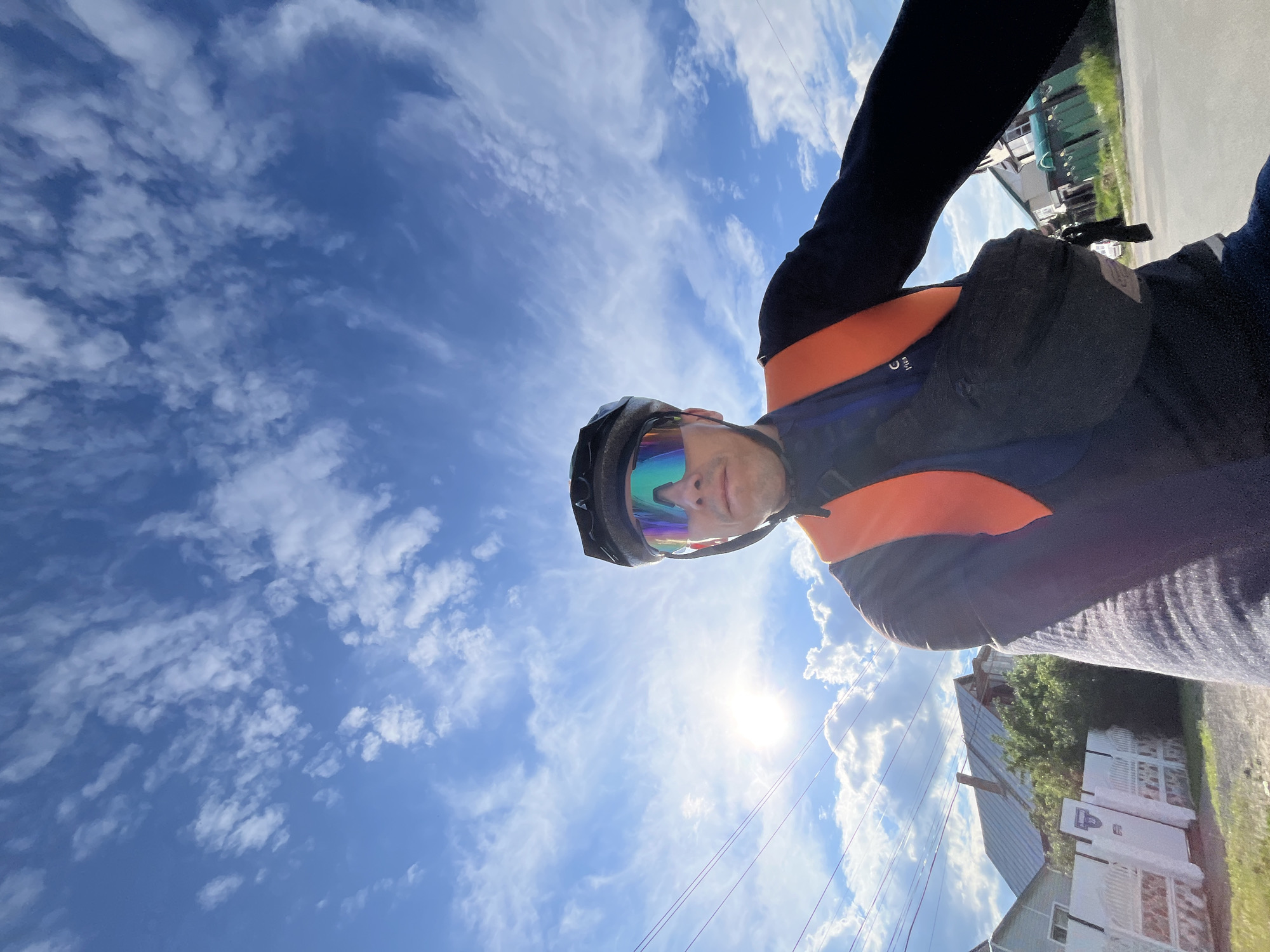The image shows a low-angle, front-facing selfie of a person wearing a black cycling helmet, colorful cycling glasses, and a dark blue long-sleeved shirt with an orange safety vest over it. A black fanny pack is worn across the chest. The person appears to be on a bicycle, looking towards the camera.
The background is a bright blue sky with scattered white, wispy clouds, indicating a sunny day. The sun is visible as a bright glare on the left side of the image, partially obscured by clouds and power lines.
In the lower left portion of the image, residential buildings with gabled roofs and white fences are visible. Green trees and shrubbery are present. Power lines run horizontally across the mid-ground. The scene suggests an outdoor setting, likely in a suburban or rural area.
The image is a selfie taken from the perspective of someone riding a bicycle, likely capturing their view of the sky and surroundings. The location is identified as Chaikovsky, Russia.